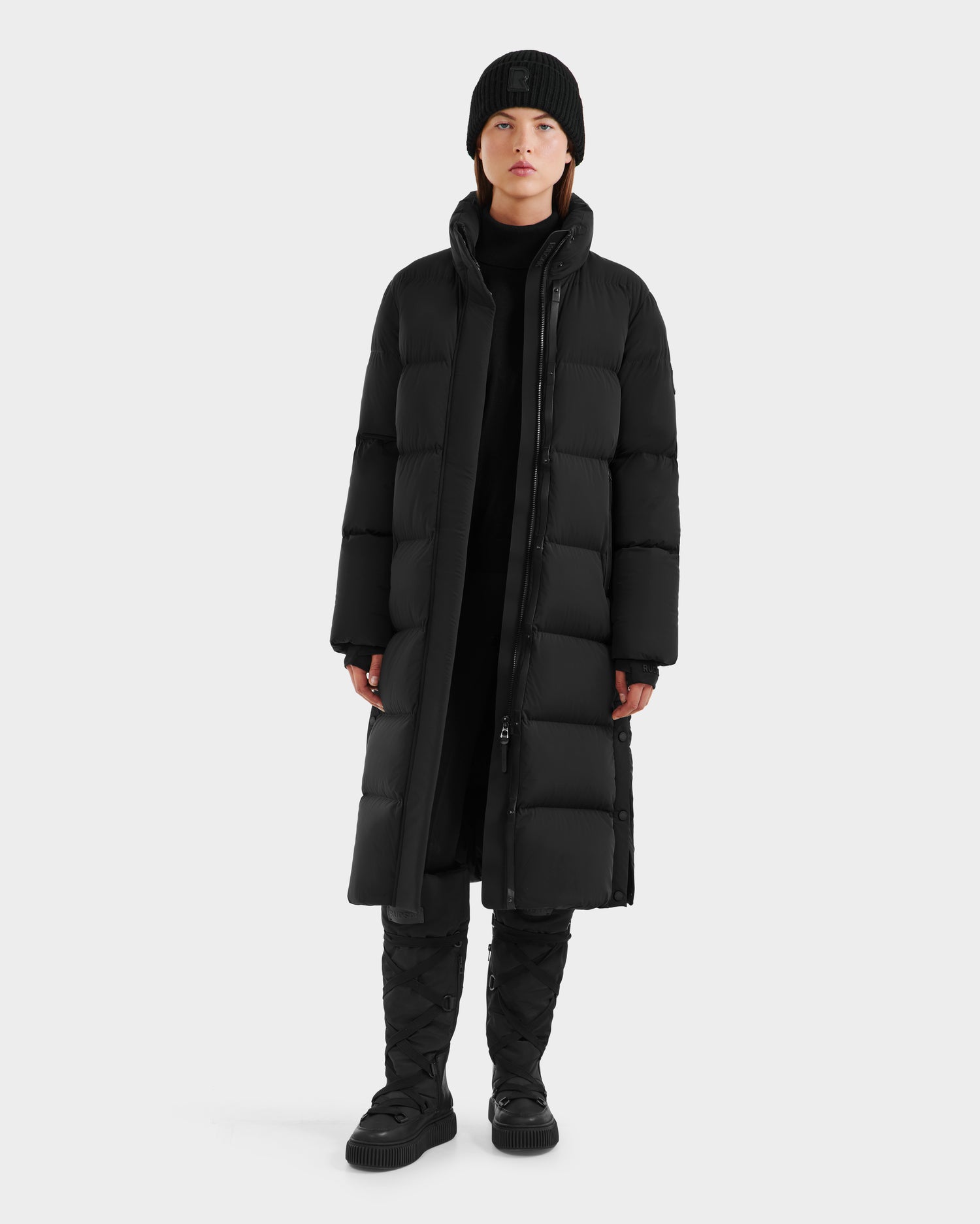 A person stands facing forward against a plain light gray background, wearing the Rudsak MAO - 8124780 Black womens long down coat, paired with a black beanie, pants, and quilted boots, with a neutral expression.