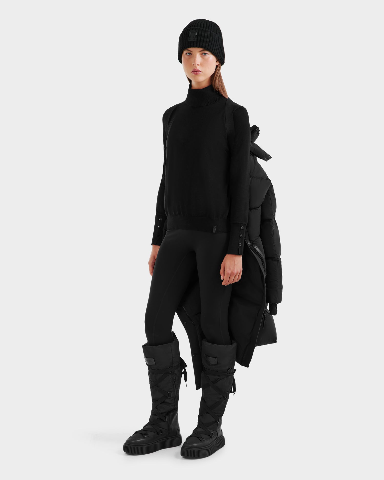 A woman wears the Rudsak MAO - 8124780 Black womens long down coat—wind and water resistant—draped over her shoulders with a knit beanie, turtleneck, leggings, and knee-high puffer boots against a plain white background.