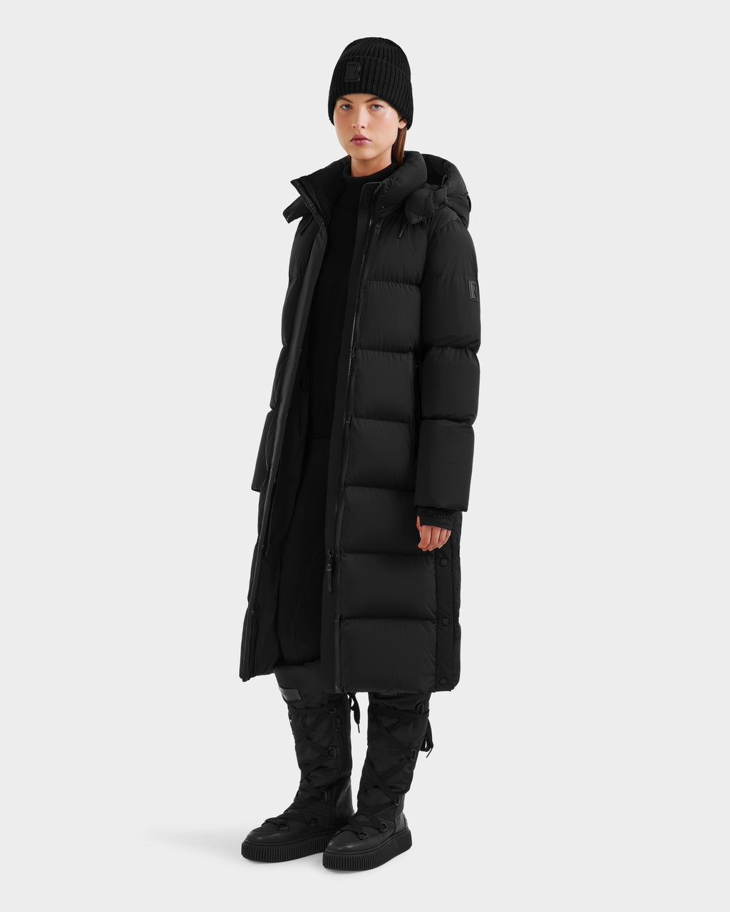 A person wearing the Rudsak MAO - 8124780 Black womens long down coat, which is wind and water resistant, paired with a black beanie, pants, and boots, stands facing forward against a plain light gray background.