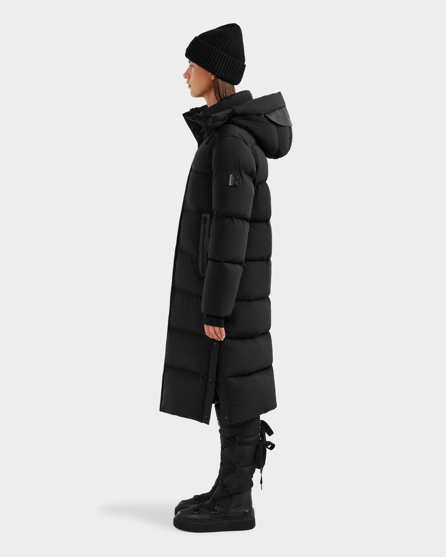 A person in profile wears the Rudsak MAO - 8124780 Black women’s long down coat with hood, paired with a black beanie and lace-up boots—wind- and water-resistant winter wear—against a plain white background.