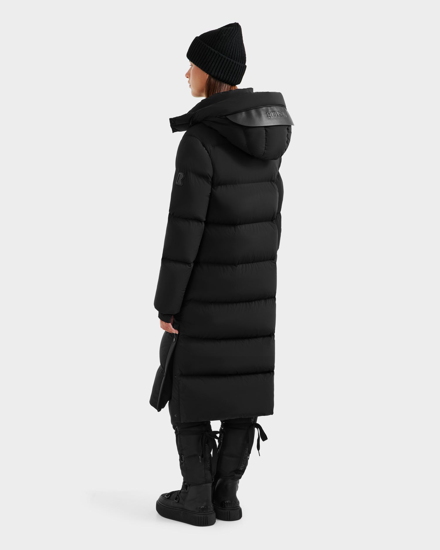A person wearing the Rudsak MAO - 8124780 Black womens long down coat, black beanie, gloves, and boots stands facing away. This wind- and water-resistant winter wear is ideal for braving the elements against a plain light background.