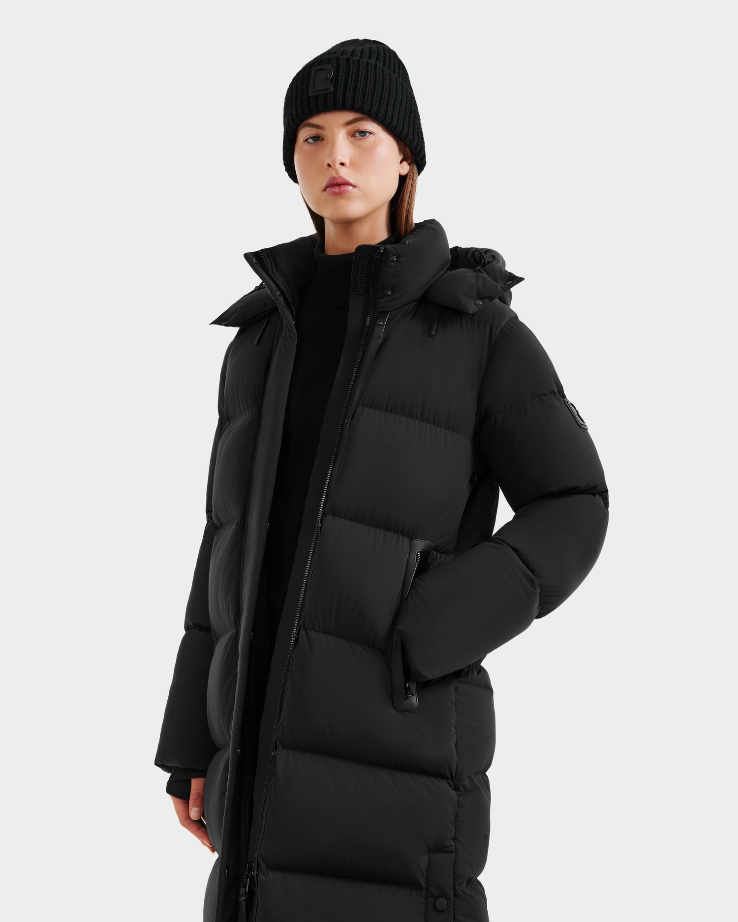 A woman in a black beanie and the Rudsak MAO - 8124780 Black women’s long quilted down coat with RDS-certified fill stands facing forward against a plain light background, one hand tucked into her pocket.