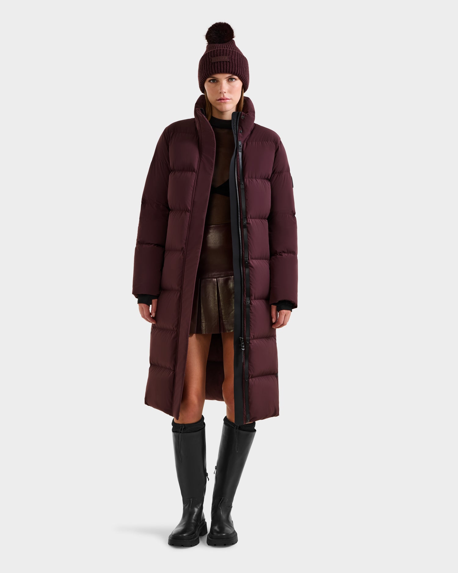 A woman wearing the Rudsak MAO - 8124780 Bordeaux wind and water resistant, RDS-certified down maroon puffer coat stands against a plain white background, styled with a matching beanie, dark brown sweater, leather skirt, and black knee-high boots.