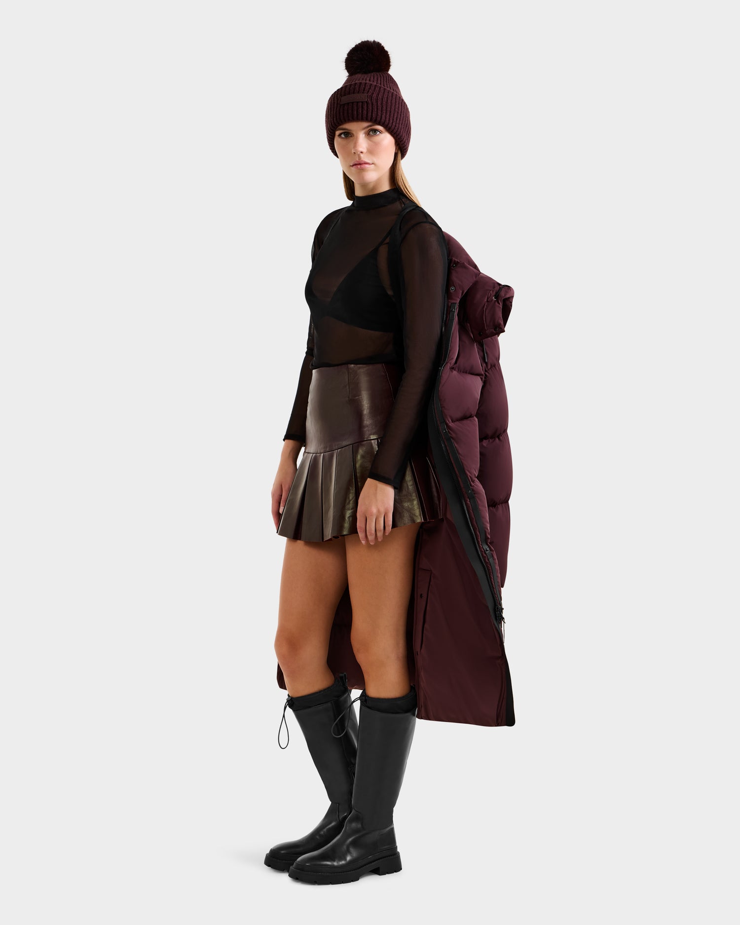 A woman stands against a light background, wearing a maroon beanie, sheer black top, metallic pleated mini skirt, black knee-high boots, and the Rudsak MAO - 8124780 Bordeaux puffer coat draped over her shoulders.