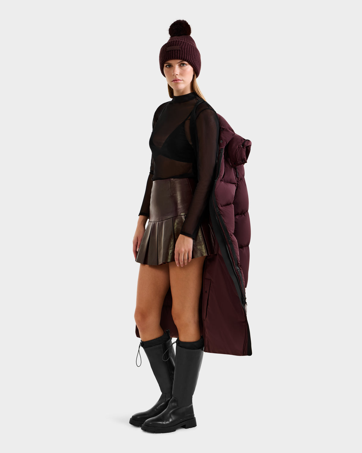A woman stands against a light background, wearing a maroon beanie, sheer black top, metallic pleated mini skirt, black knee-high boots, and the Rudsak MAO - 8124780 Bordeaux puffer coat draped over her shoulders.