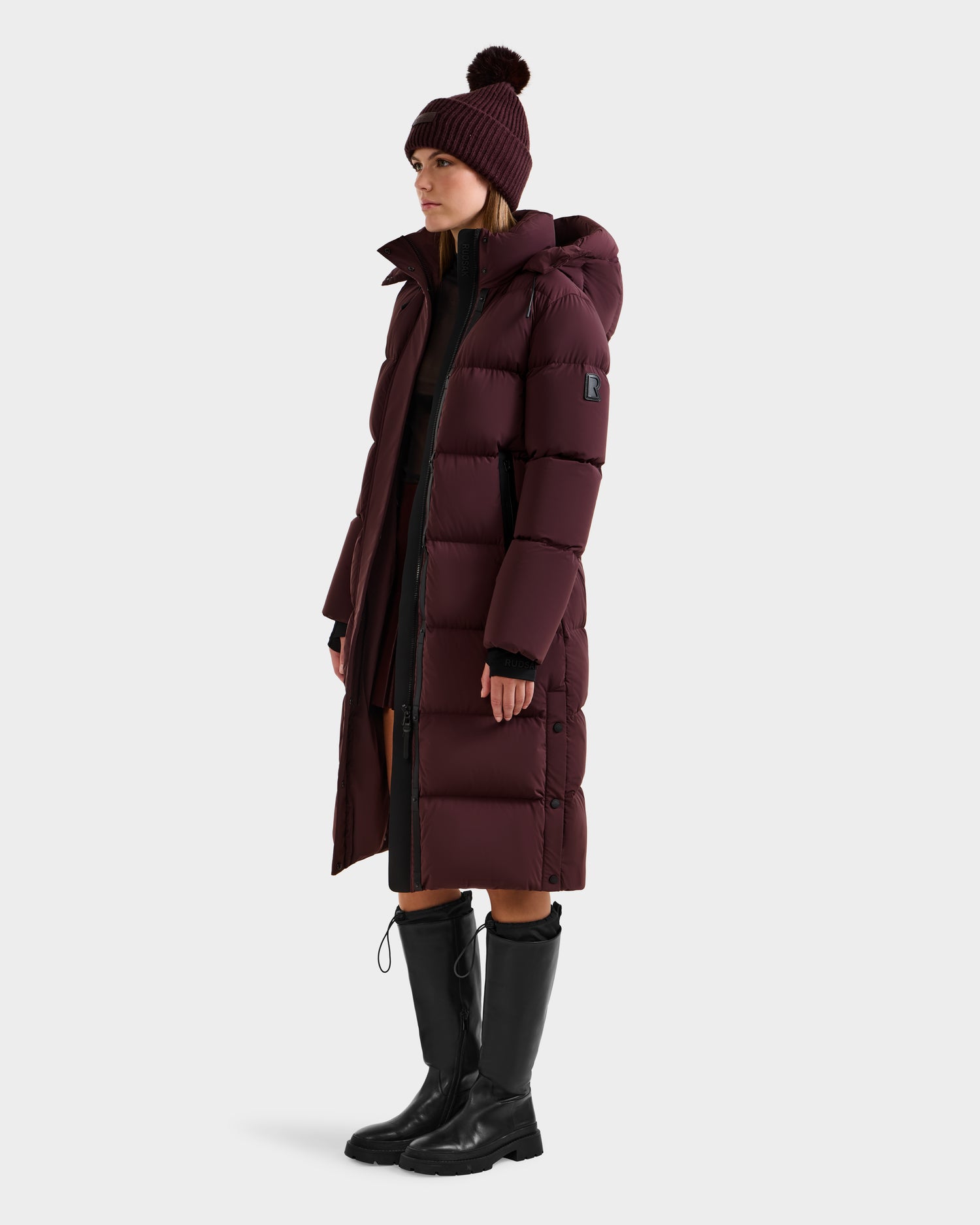 A woman wearing the Rudsak MAO - 8124780 Bordeaux women's long quilted down coat stands against a white background; the wind- and water-resistant coat with RDS-certified down is styled with a matching pom-pom beanie, black clothes, and knee-high boots.