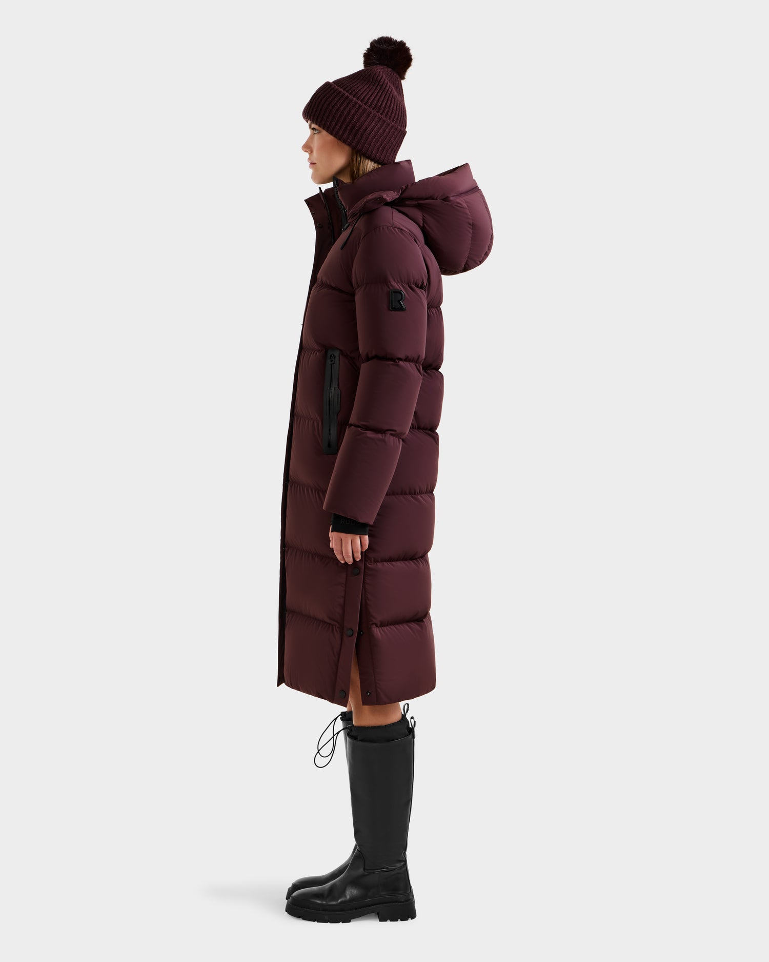 A woman in profile wears the Rudsak MAO - 8124780 Bordeaux women's long quilted down coat with a hood, wind and water resistant, paired with a matching maroon pom-pom hat and black knee-high boots against a plain light background.