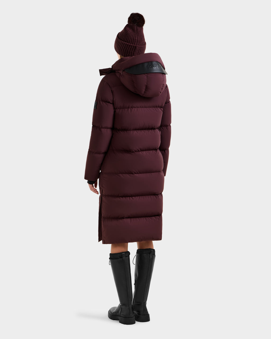 A person wearing the Rudsak MAO - 8124780 Bordeaux women's long quilted down coat with hood, wind and water resistant, paired with a knit beanie and tall black boots, stands facing away against a plain white background.