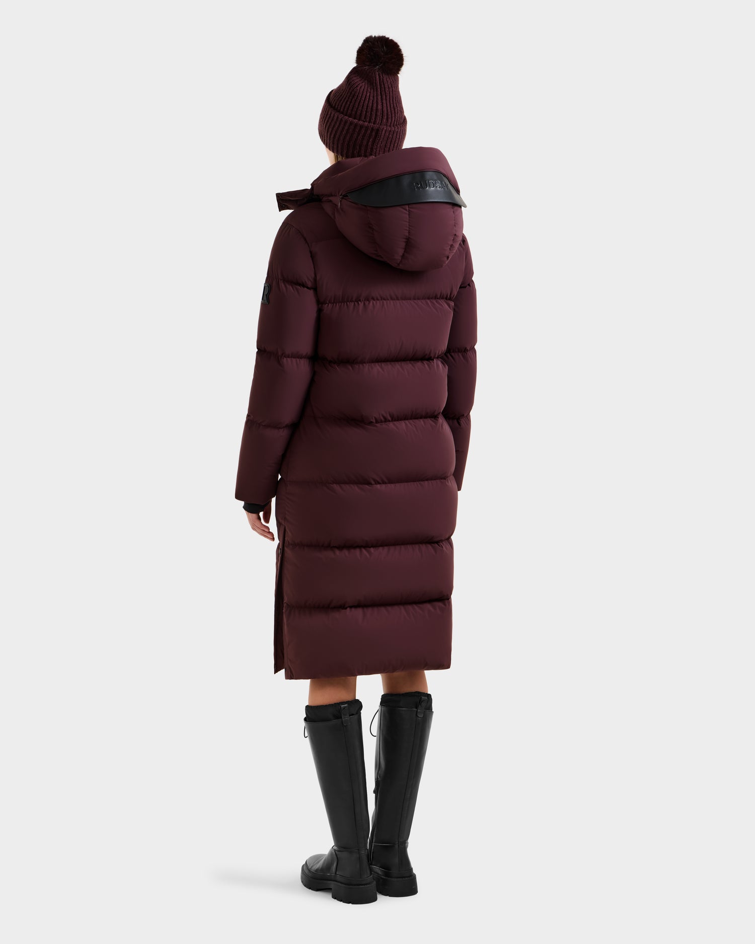A person wearing the Rudsak MAO - 8124780 Bordeaux women's long quilted down coat with hood, wind and water resistant, paired with a knit beanie and tall black boots, stands facing away against a plain white background.