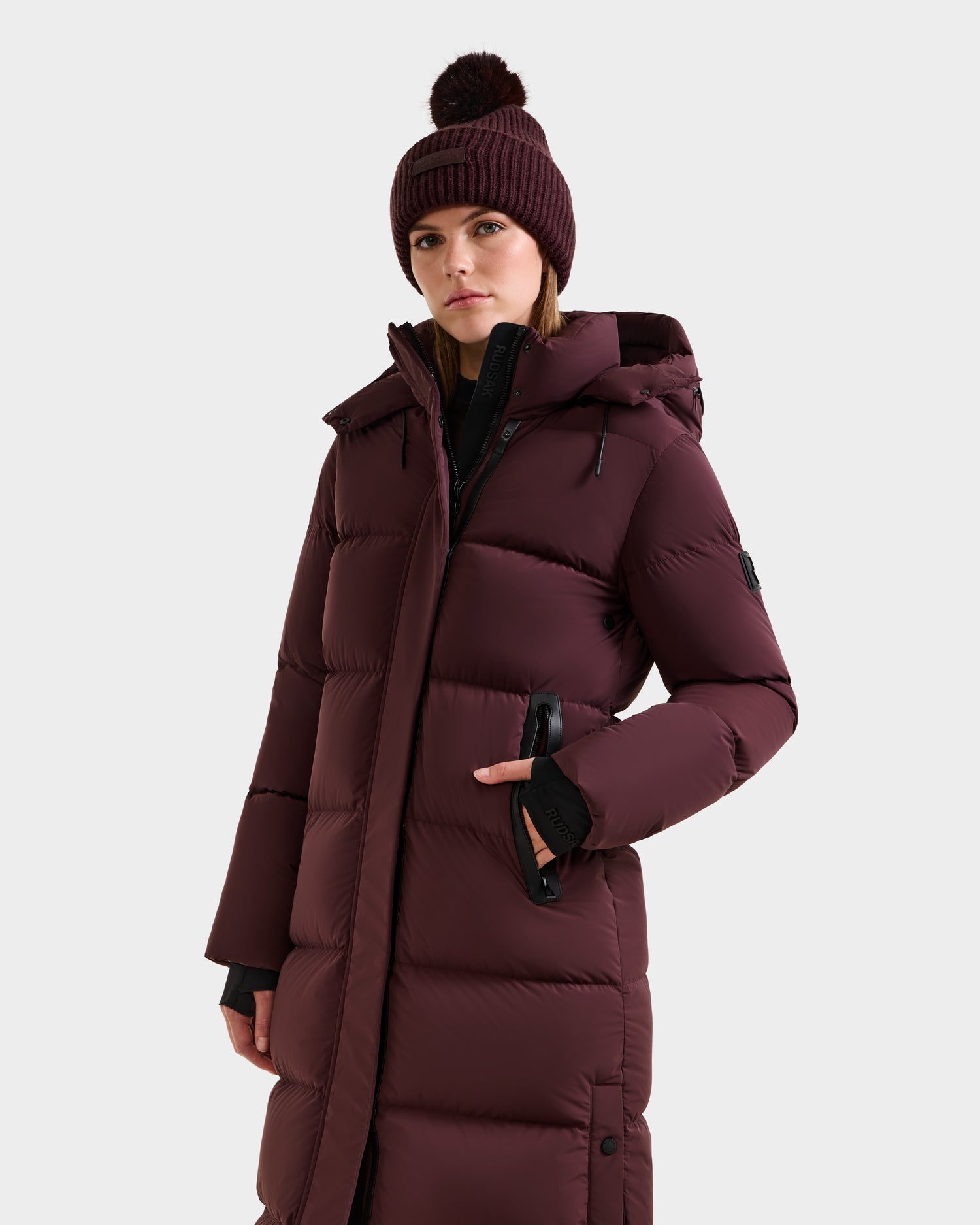 A person in the Rudsak MAO - 8124780 Bordeaux women's long quilted down coat with a hood and a matching knit beanie stands against a plain light background. The coat features wind- and water-resistant, RDS-certified down.