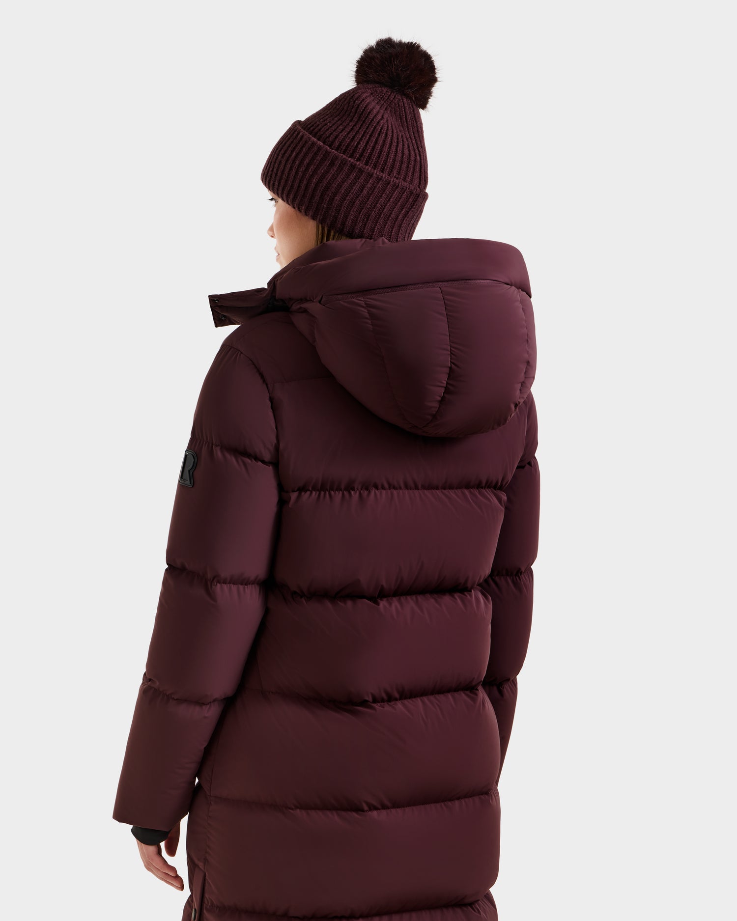 A person wearing the Rudsak MAO - 8124780 Bordeaux women's long quilted down coat with hood, wind and water resistant, RDS-certified down, and a matching pom-pom beanie, seen from the back against a plain light background.