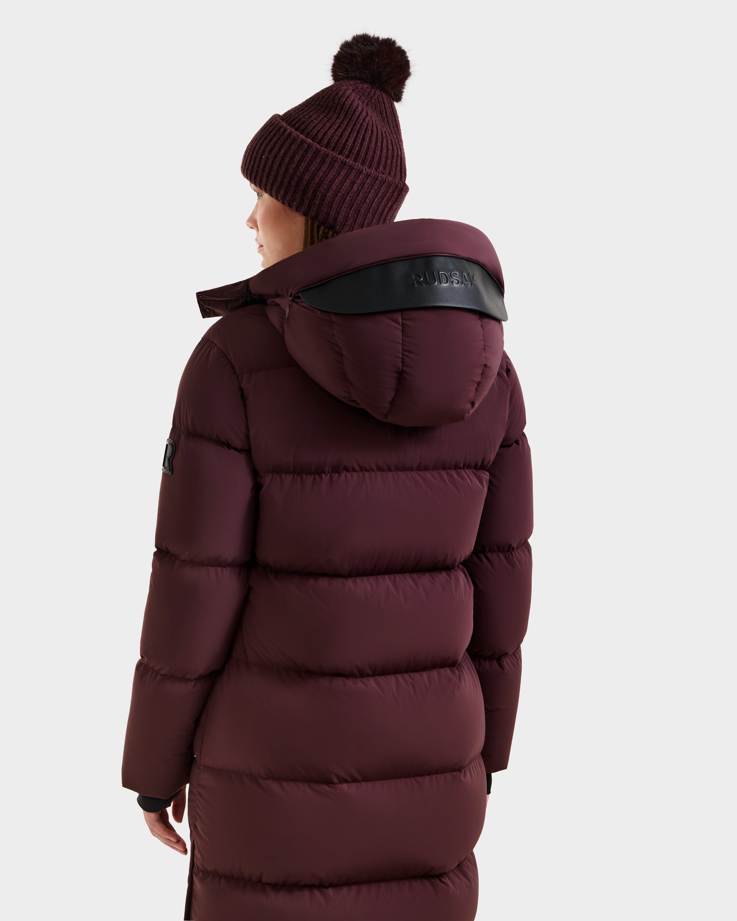 A person wearing the Rudsak MAO - 8124780 Bordeaux women's long quilted down coat with a large hood and a matching knit beanie, seen from behind against a plain white background.