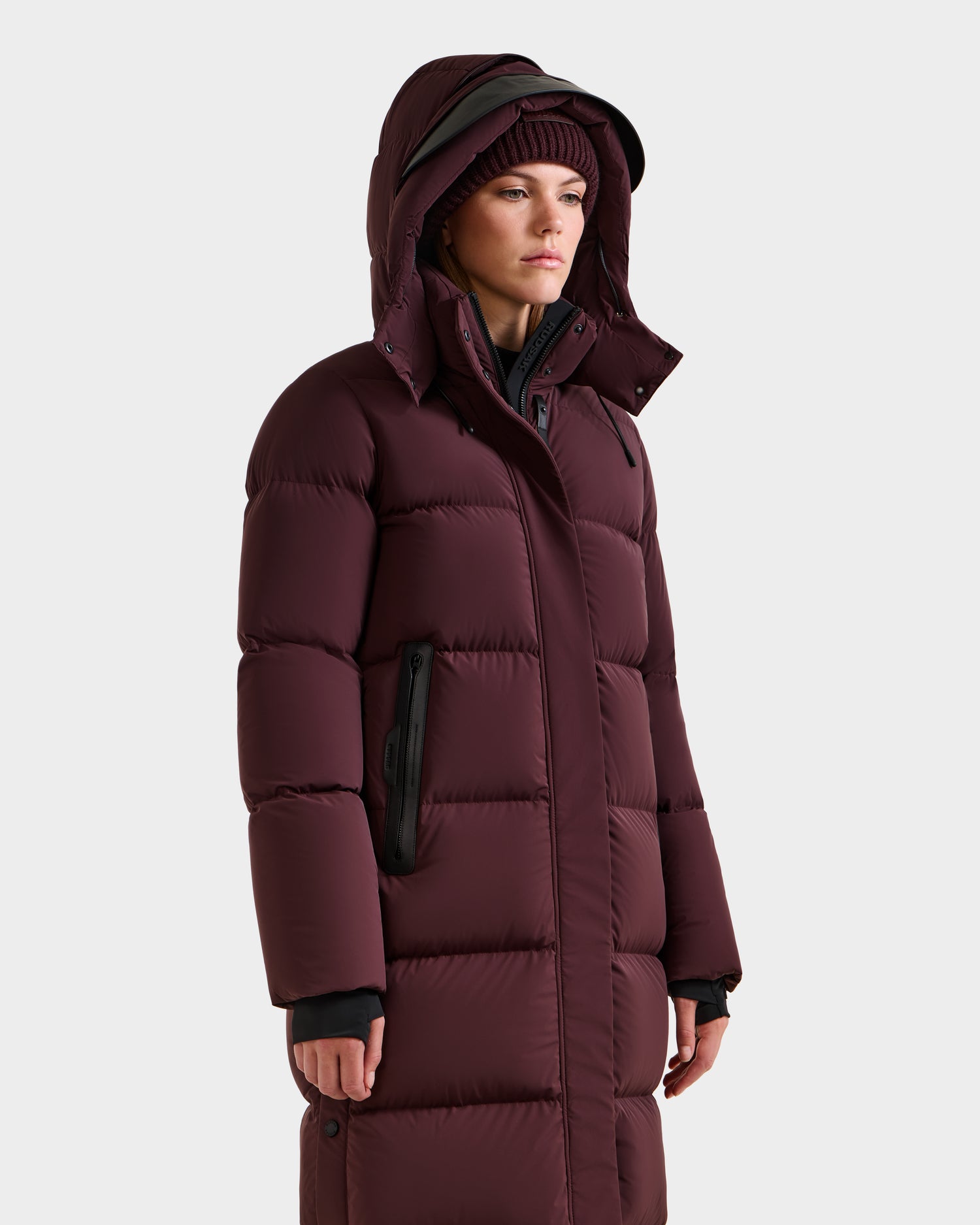 A person wears the Rudsak MAO - 8124780 Bordeaux women’s long quilted down coat with hood, wind and water resistant, and a matching knit beanie, standing against a light background, looking slightly to the side.