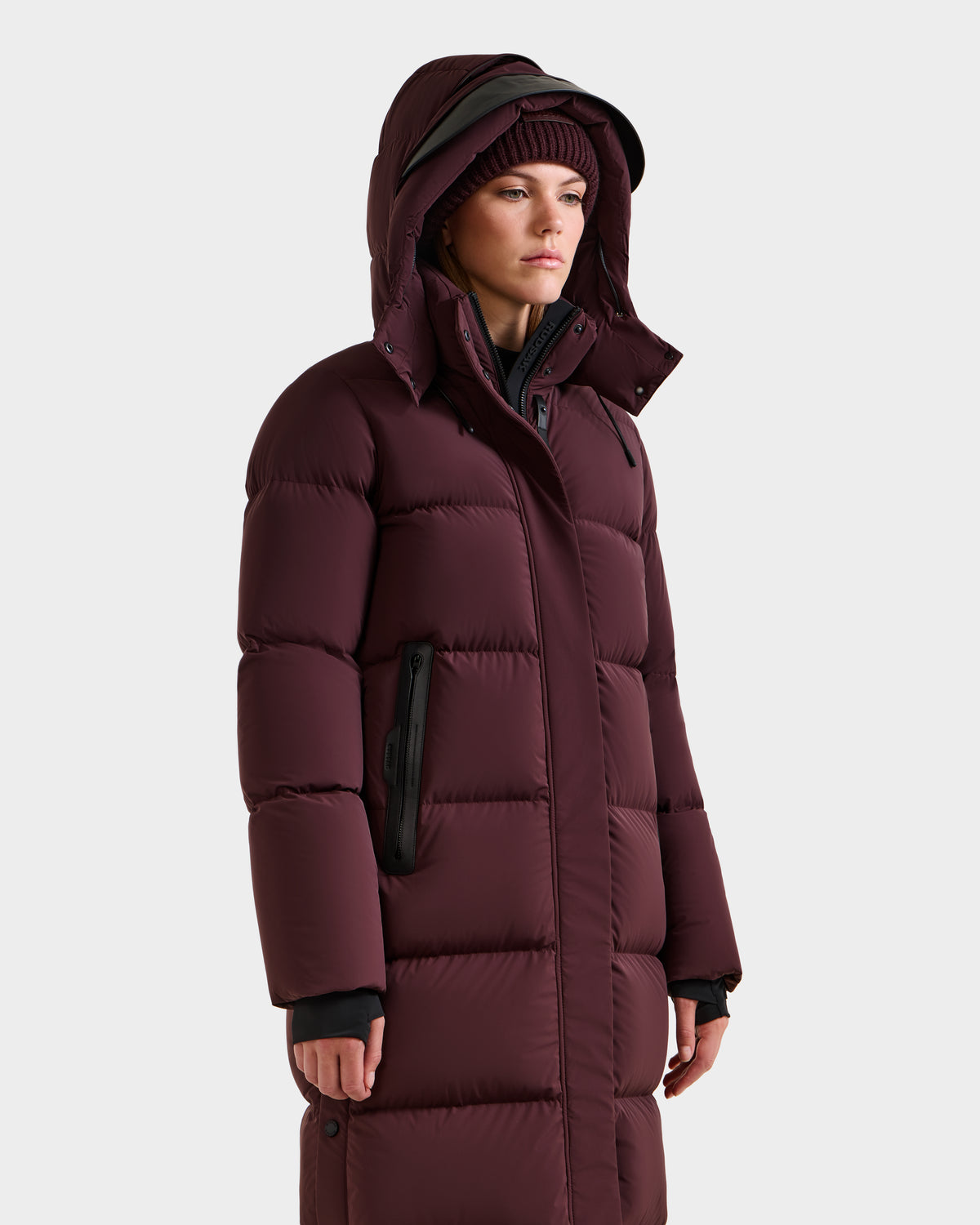 A person wears the Rudsak MAO - 8124780 Bordeaux women’s long quilted down coat with hood, wind and water resistant, and a matching knit beanie, standing against a light background, looking slightly to the side.