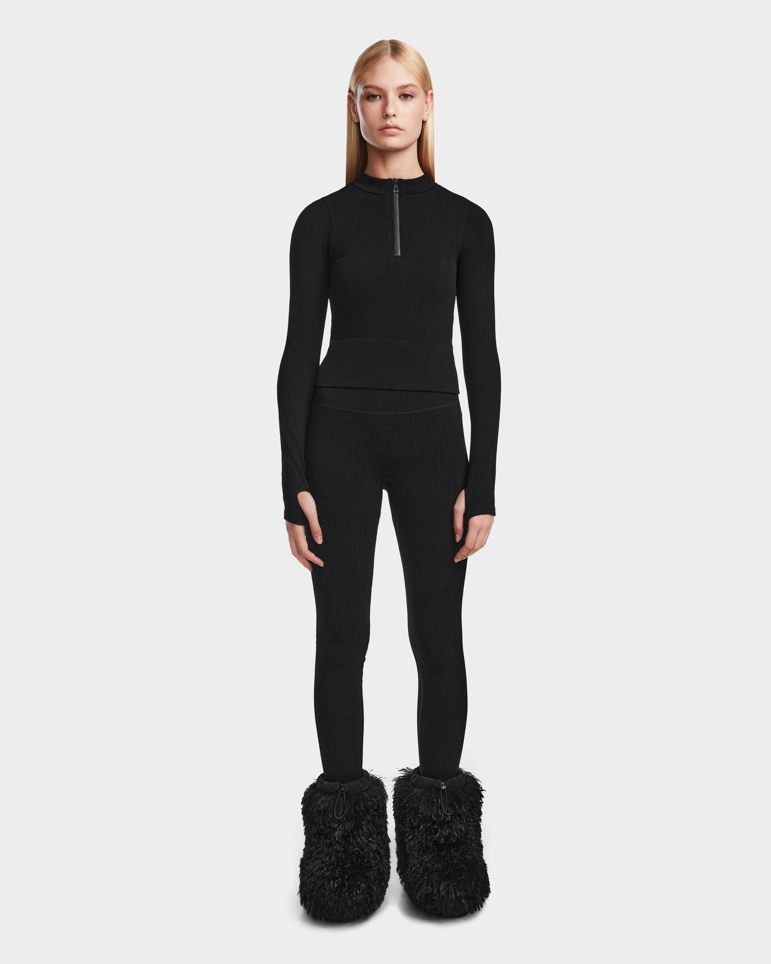 A person stands facing forward, wearing Rudsak’s MARIAN - 8524513 Monogram Print Black fitted stretch top with a zipper, matching black leggings, and large fuzzy slippers—a chic choice for any winter wardrobe. The background is plain and light-colored.