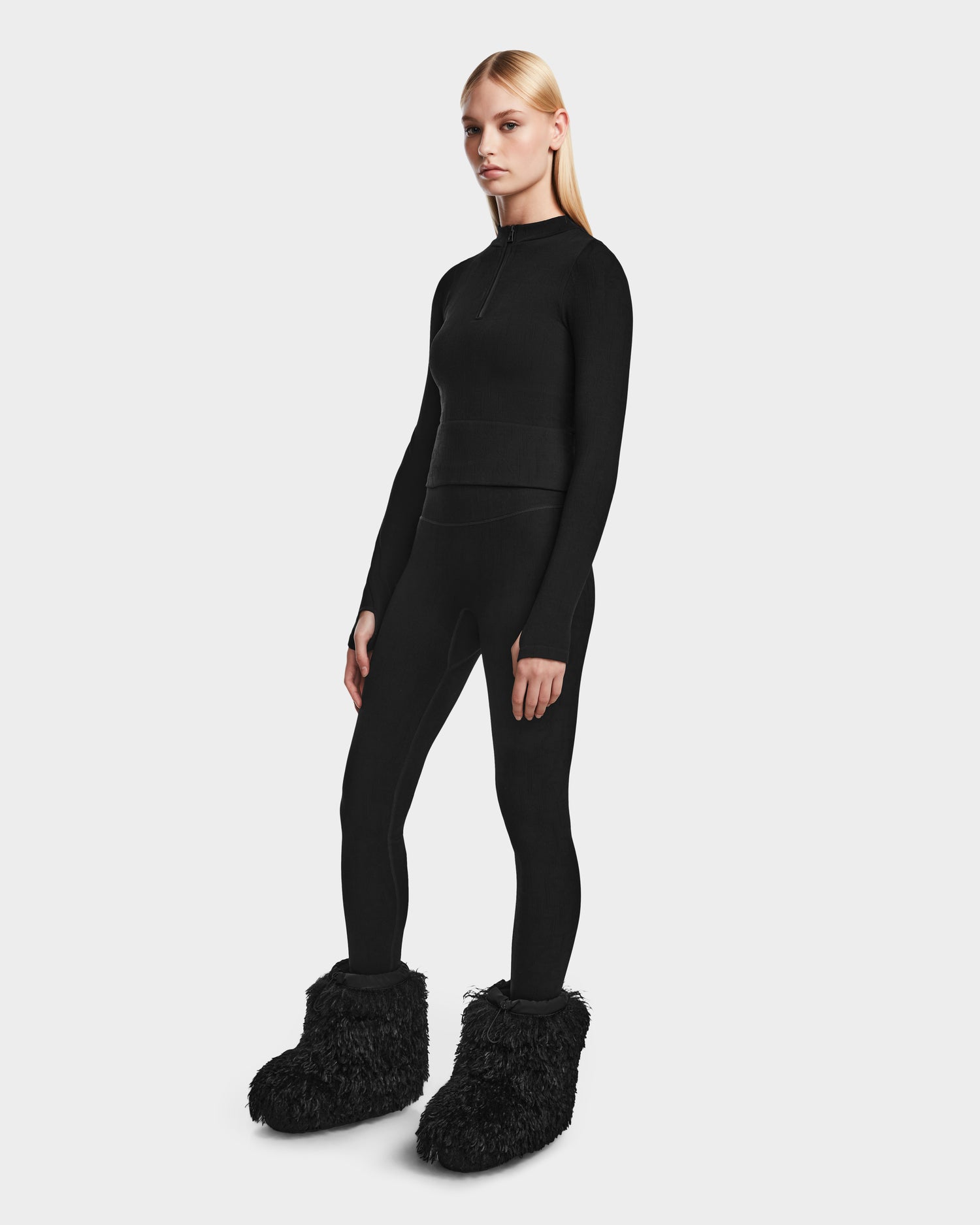 A woman in Rudsak’s MARIAN - 8524513 Monogram Print Black stretch top and leggings with furry black slippers stands against a white backdrop—an effortless après-ski look for winter.