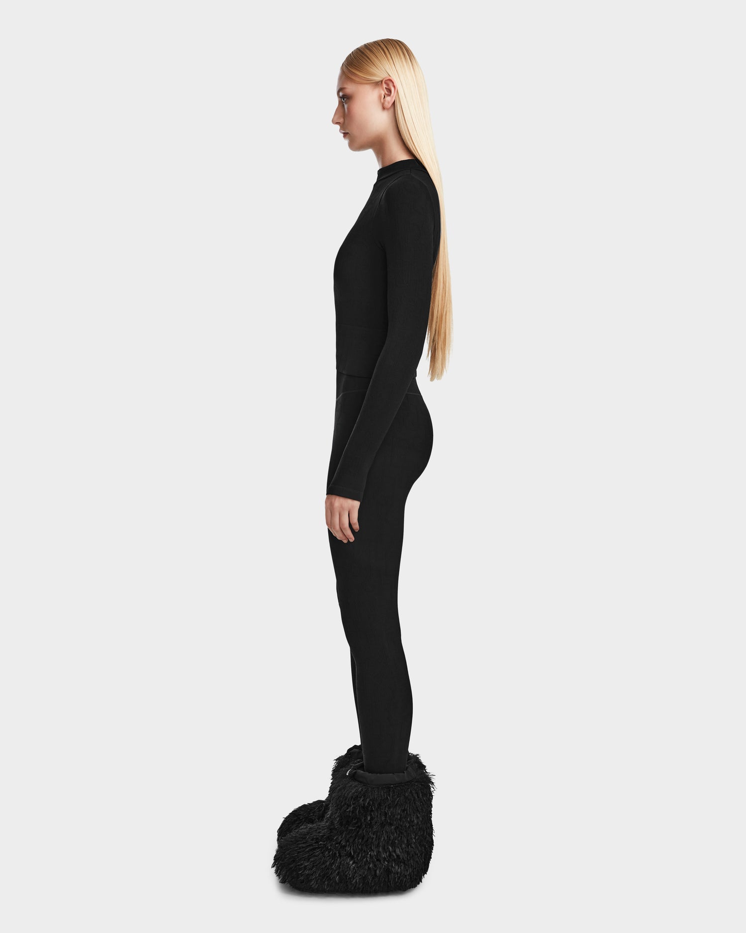 A woman with long blonde hair stands in profile, wearing the Rudsak MARIAN - 8524513 Monogram Print Black stretch top and large, fluffy black footwear, perfect for après-ski style against a plain light gray background.