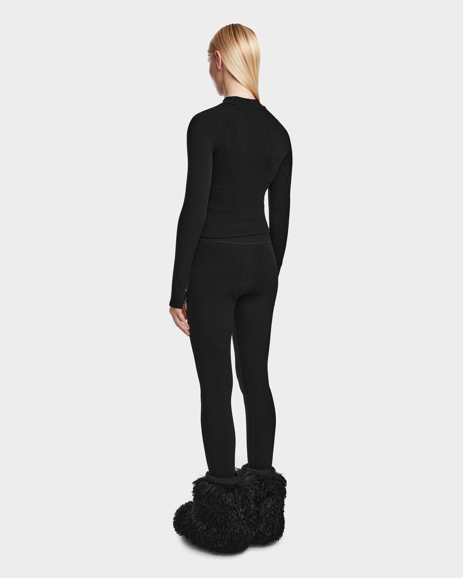 A person with long blonde hair faces away, modeling Rudsaks MARIAN - 8524513 Monogram Print Black fitted stretch top and black leggings, paired with large furry boots, set against a plain light gray background.