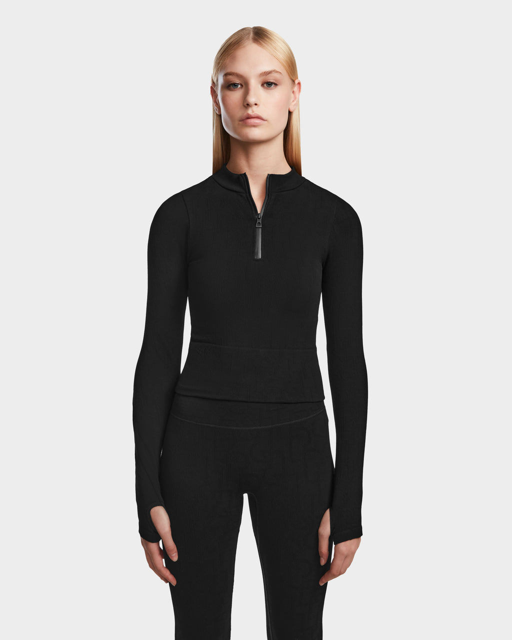 A woman with long blonde hair wears the Rudsak MARIAN - 8524513 Monogram Print Black top and matching black pants, standing against a light gray background—a sleek choice for any winter wardrobe.