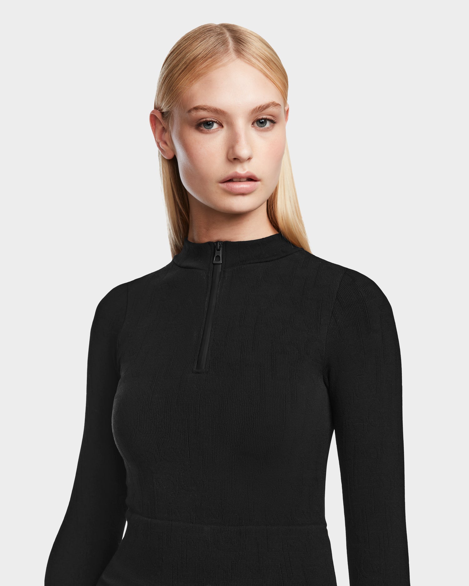 A woman with straight blonde hair wears the Rudsak MARIAN - 8524513 Monogram Print Black stretch top with a short front zipper, standing against a light background—an ideal addition to your winter wardrobe.