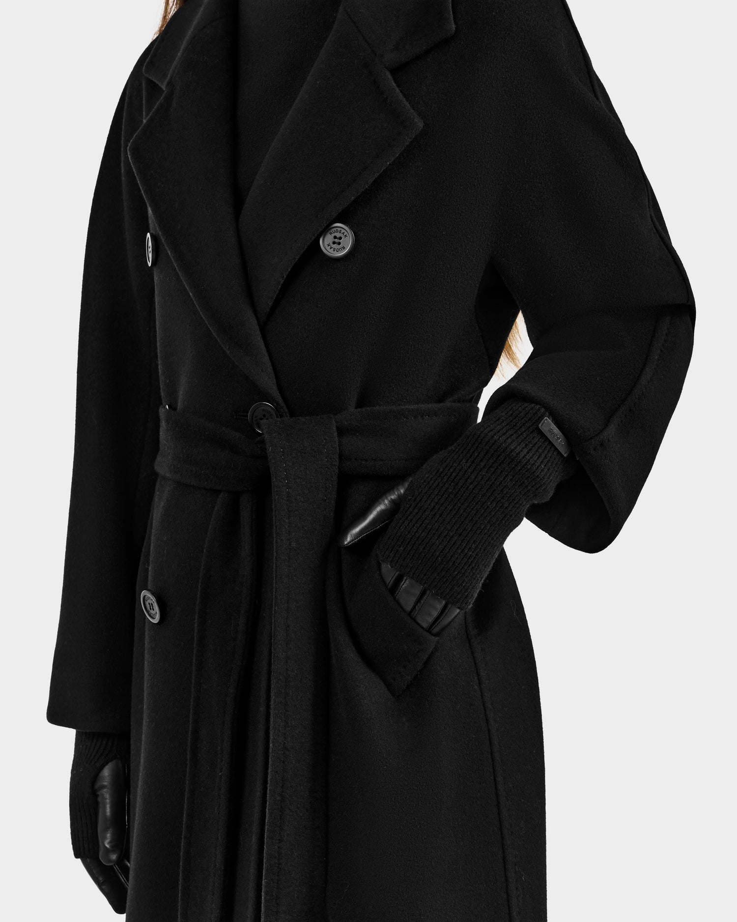 A person in a long black belted coat with large lapels, a black turtleneck sweater, and Rudsak MARINE - 8424516 Black women's leather gloves. Their face is not visible in the image.