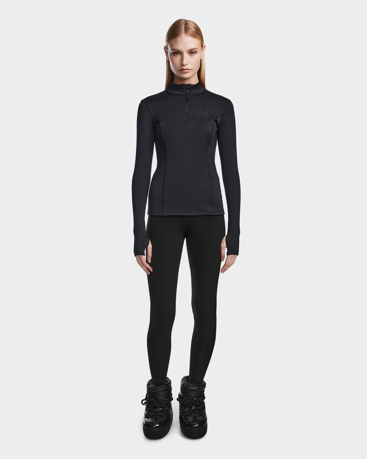 A woman with straight blonde hair stands facing forward, wearing Rudsak’s MARIS - 8524603 Black fitted women’s active top and black leggings—ideal ski layering for winter sports—against a plain white background.