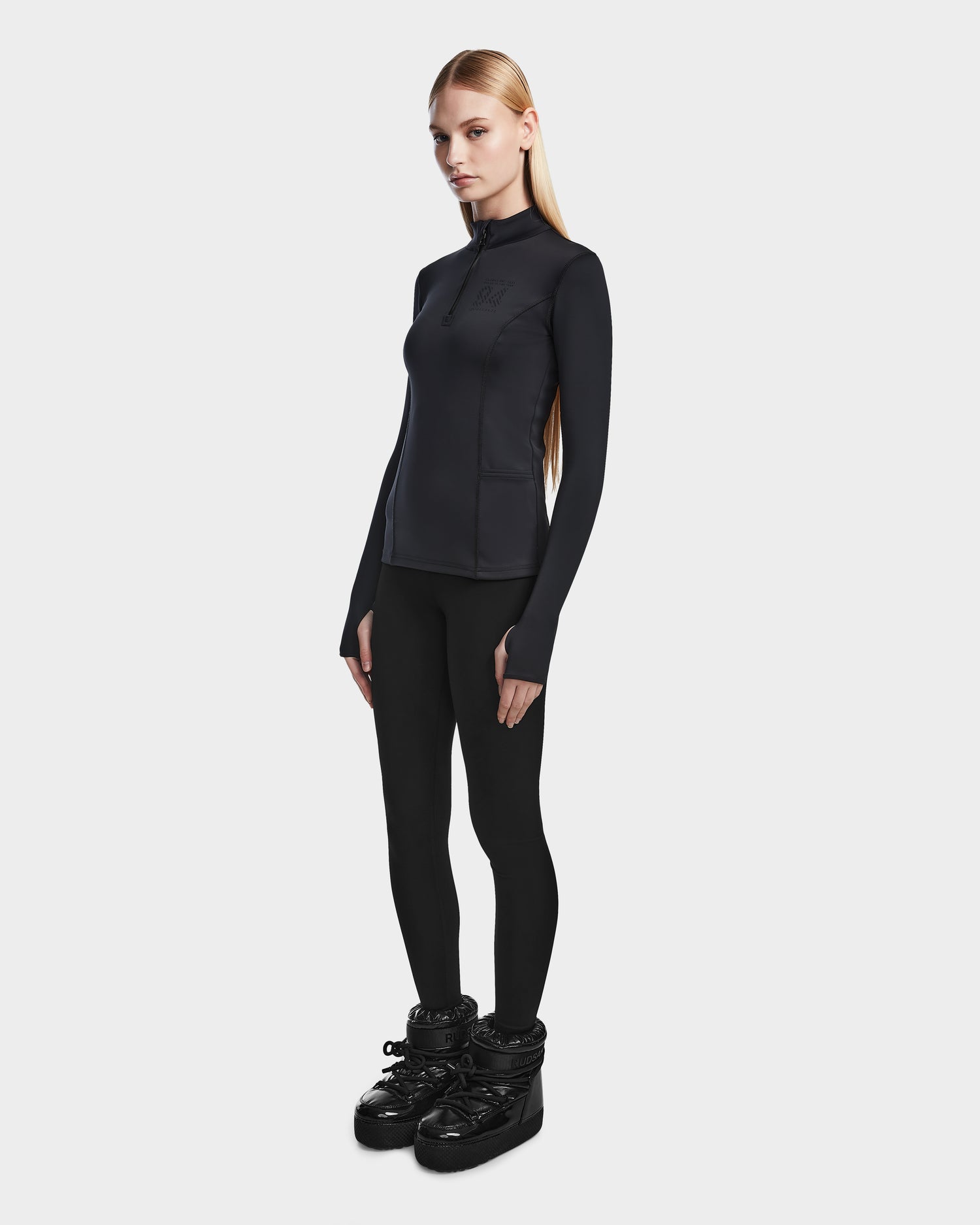 A woman with straight blonde hair and a neutral expression stands against a white background, wearing the Rudsak MARIS - 8524603 Black womens active top, black leggings, and shiny black snow boots—an ideal ski layering look.