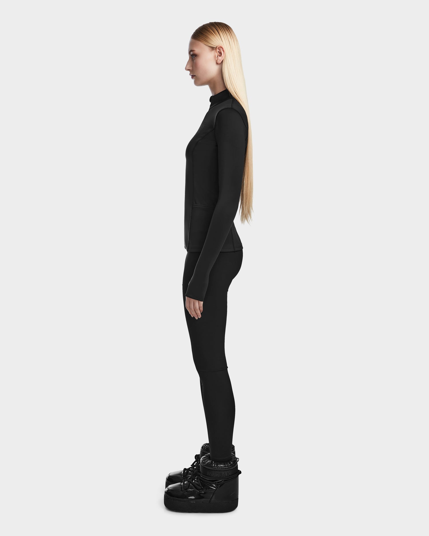 A woman with long straight blonde hair stands in profile against a white background, wearing the fitted Rudsak MARIS - 8524603 Black womens active top, black leggings, and shiny black boots.