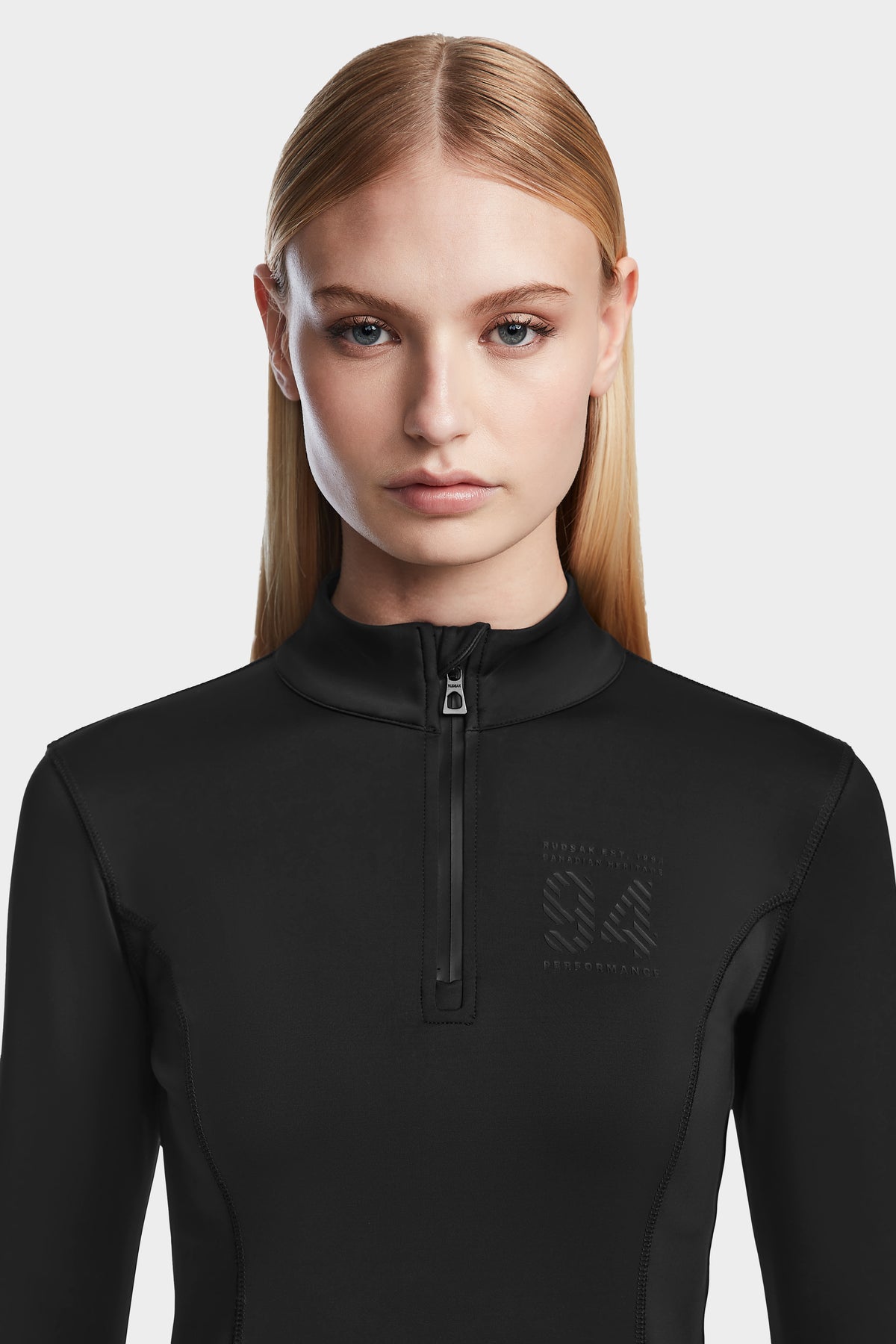 A woman with straight blonde hair wears the Rudsak MARIS - 8524603 Black women’s active top, ideal for ski layering, and looks directly at the camera against a plain light background.
