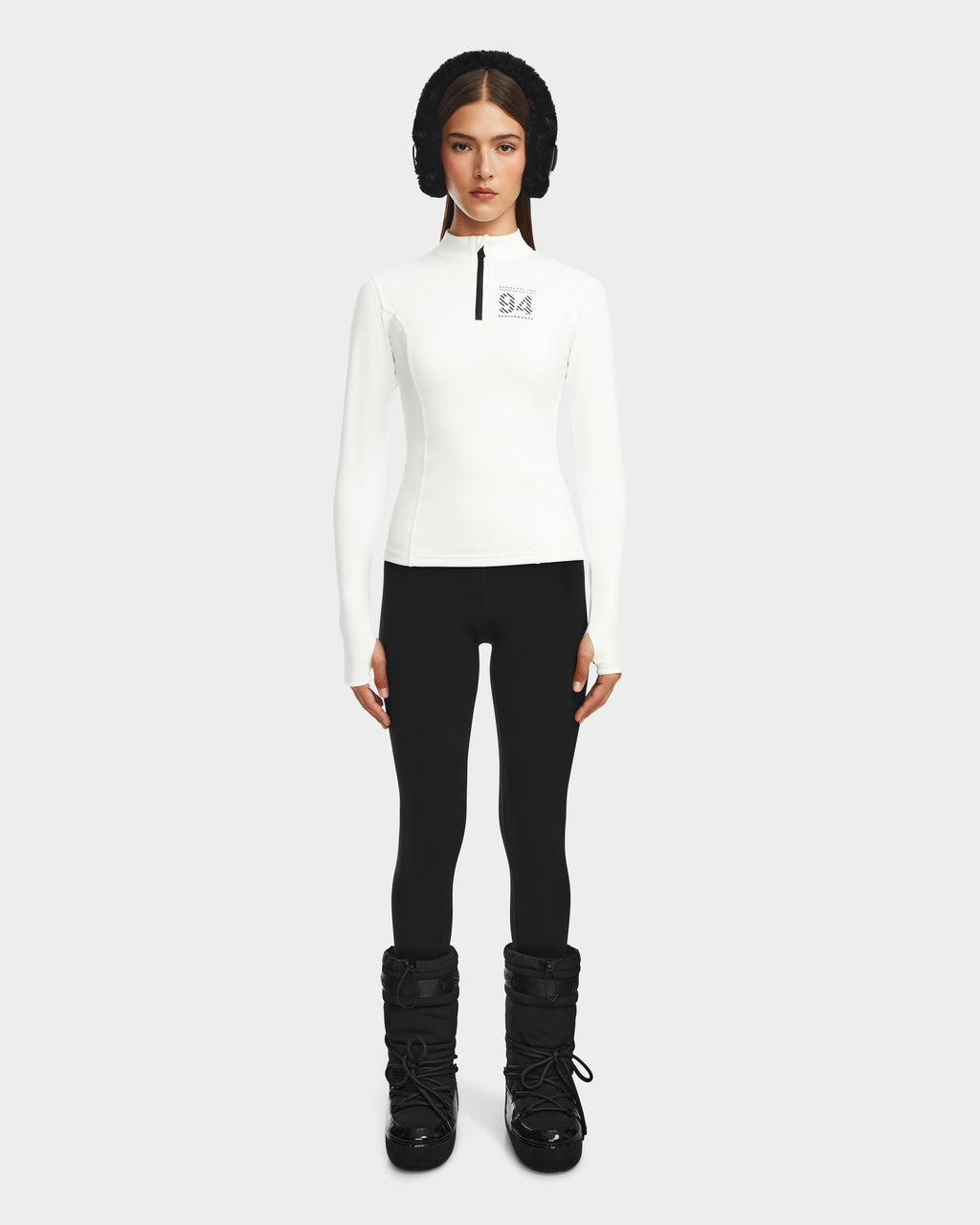 A woman stands against a plain background wearing the Rudsak MARIS - 8524603 Optic White womens active top, black leggings, black winter boots, and a black padded headband—a sleek look for ski layering or winter sports. She looks forward with her arms at her sides.