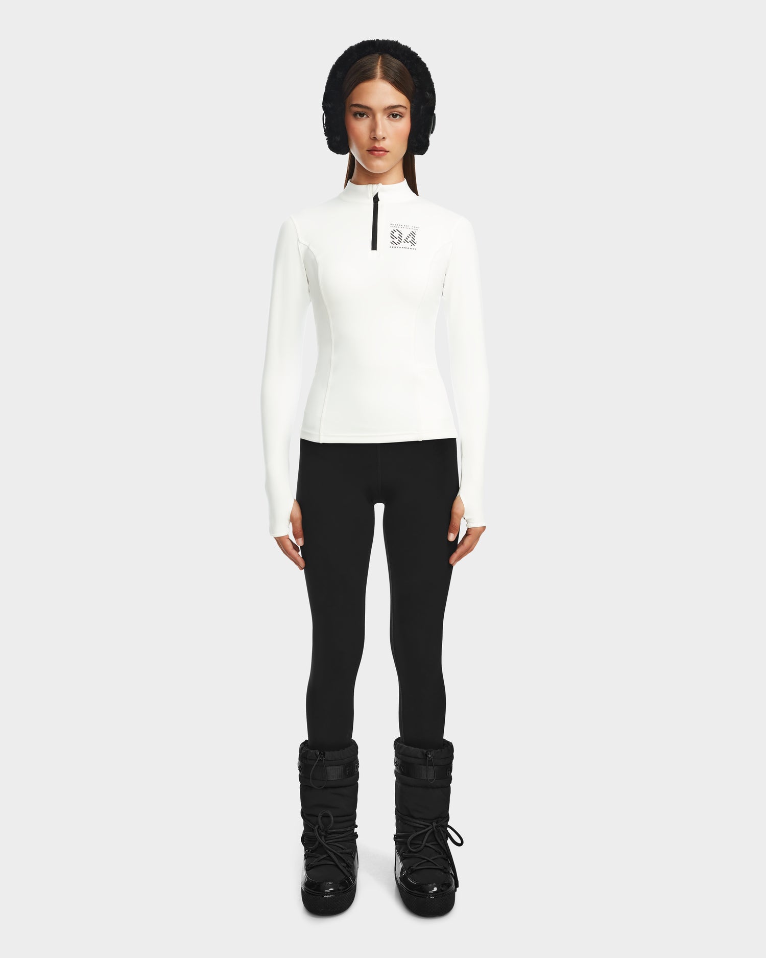 A woman stands against a plain background wearing the Rudsak MARIS - 8524603 Optic White womens active top, black leggings, black winter boots, and a black padded headband—a sleek look for ski layering or winter sports. She looks forward with her arms at her sides.