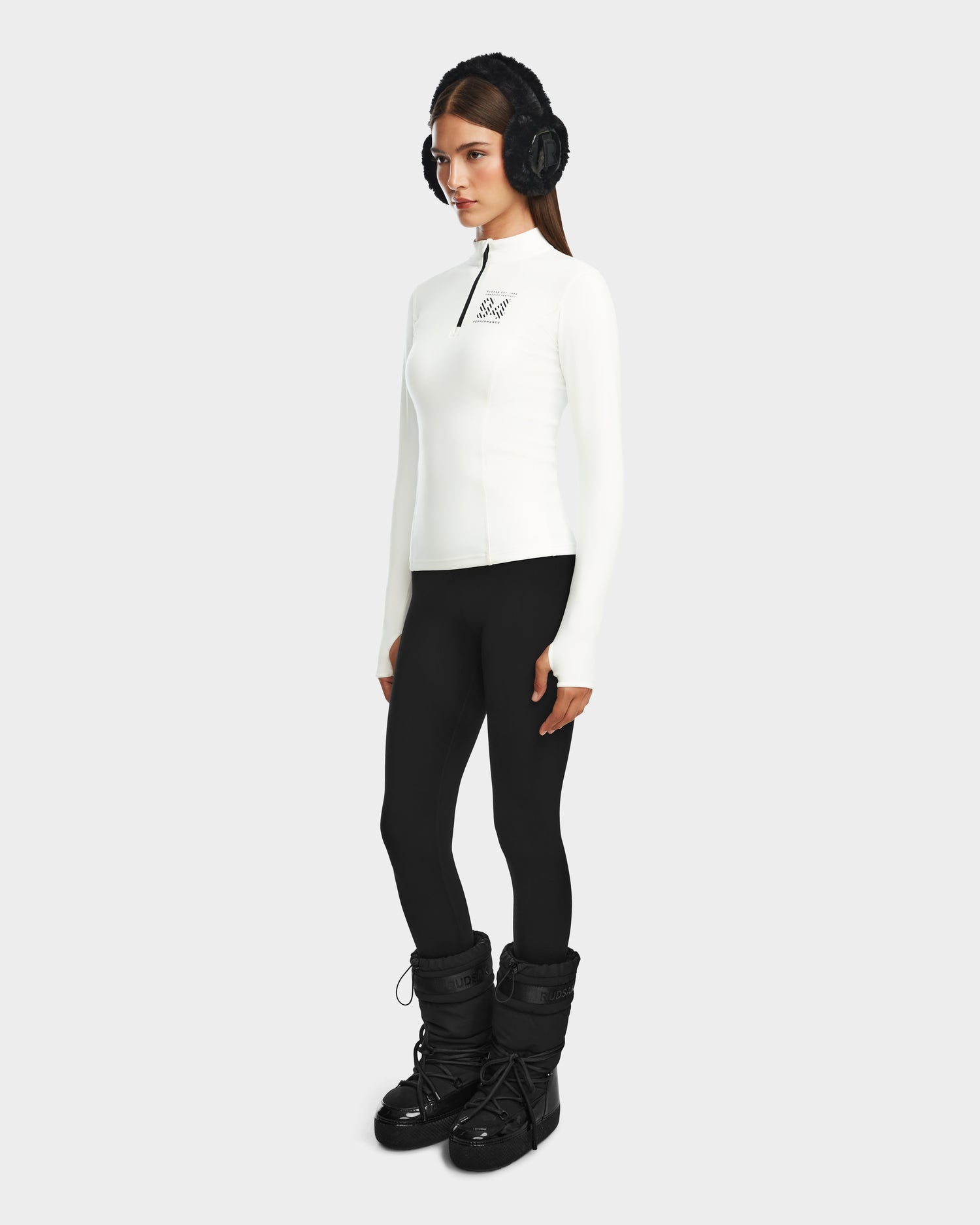 A woman with long brown hair and a neutral expression stands against a plain background, wearing Rudsaks MARIS - 8524603 Optic White fitted zip-up women’s active top, paired with black earmuffs, black leggings, and winter boots—ideal for ski layering.