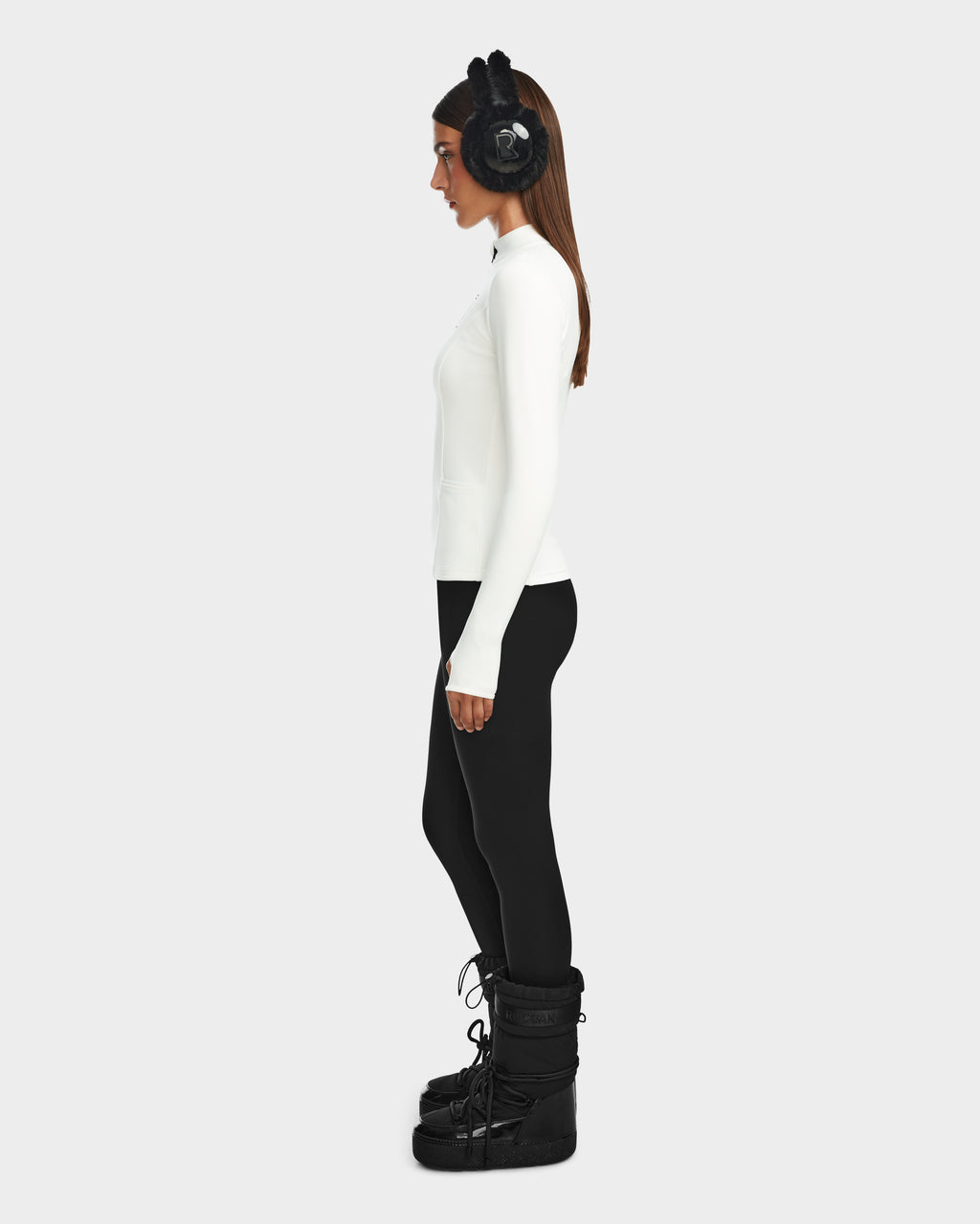 A woman stands in profile against a plain background, wearing Rudsak’s MARIS - 8524603 Optic White women’s active top, perfect for ski layering, paired with black earmuffs, leggings, and winter boots.