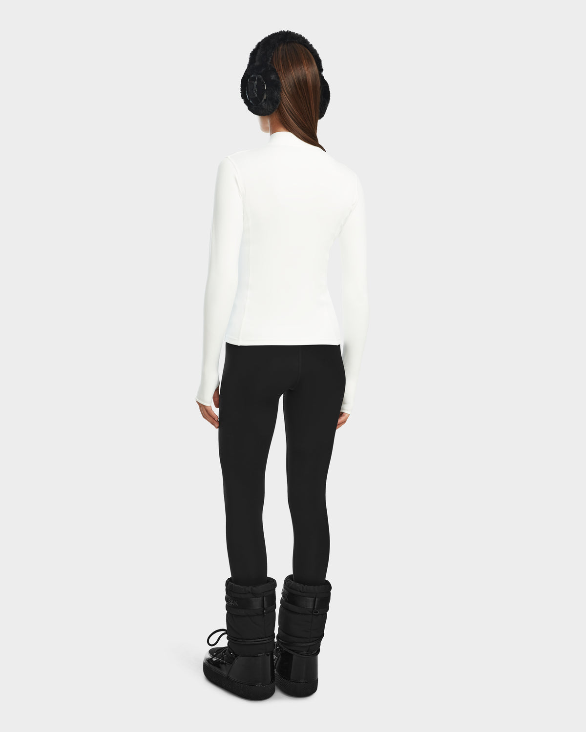 A woman faces away, wearing Rudsaks MARIS - 8524603 Optic White active top with black earmuffs, leggings, and snow boots—ideal for ski layering or winter sports against a plain light background.