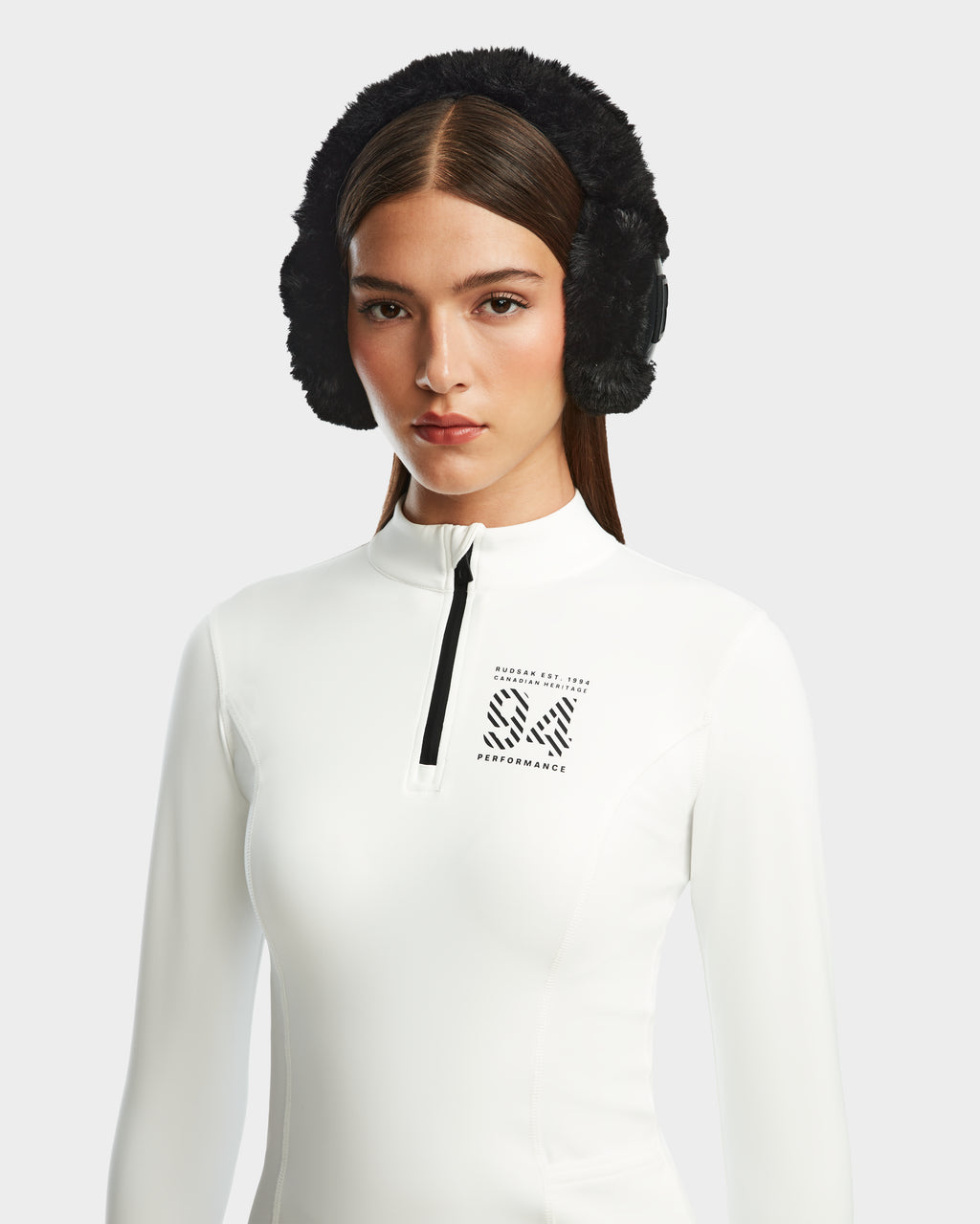 A woman with straight brown hair wears black furry earmuffs and the Rudsak MARIS - 8524603 Optic White women’s active top, featuring “Ski Performance” on the chest, perfect for ski layering, against a plain light background.