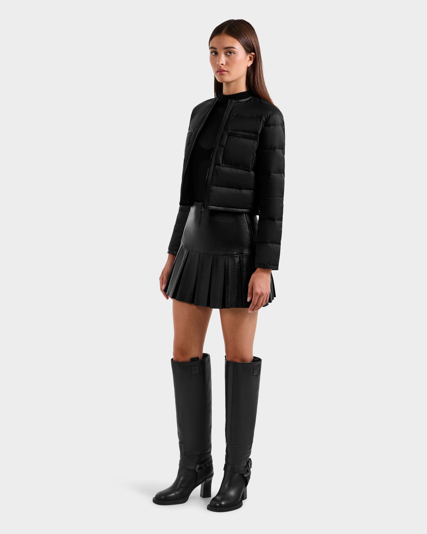 A woman models the Rudsak MARKLE - 8125033 Black cropped down jacket with vegan leather trim, paired with a black top, mini pleated skirt, and knee-high boots against a plain light gray background.