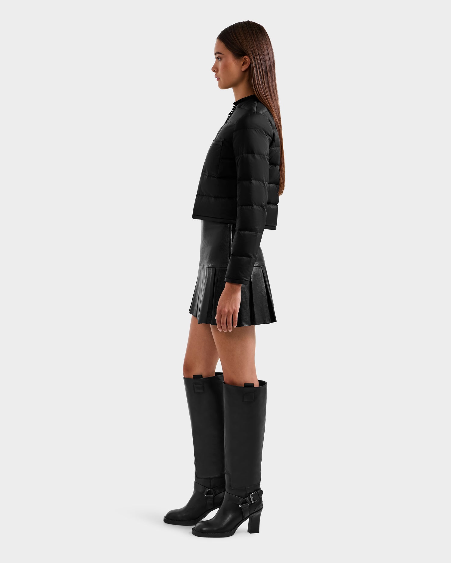 A woman stands in profile against a plain light background, wearing the Rudsak MARKLE - 8125033 Black lightweight down jacket with vegan leather trim, styled with a black pleated mini skirt and knee-high black heeled boots.