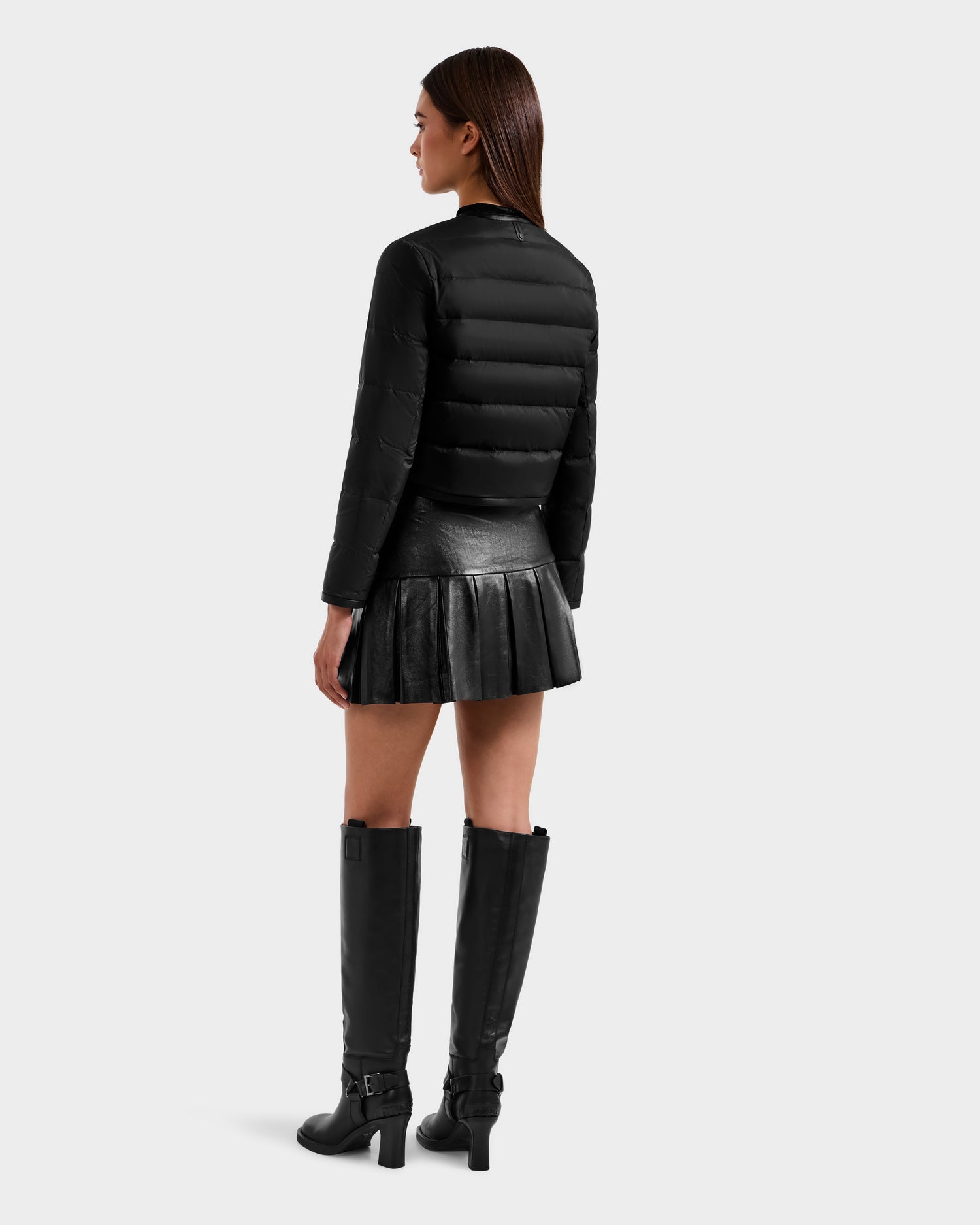 A woman with straight brown hair stands against a light gray background, wearing the Rudsak MARKLE - 8125033 Black women’s cropped down jacket with vegan leather trim, paired with a black pleated skirt and knee-high buckle boots.