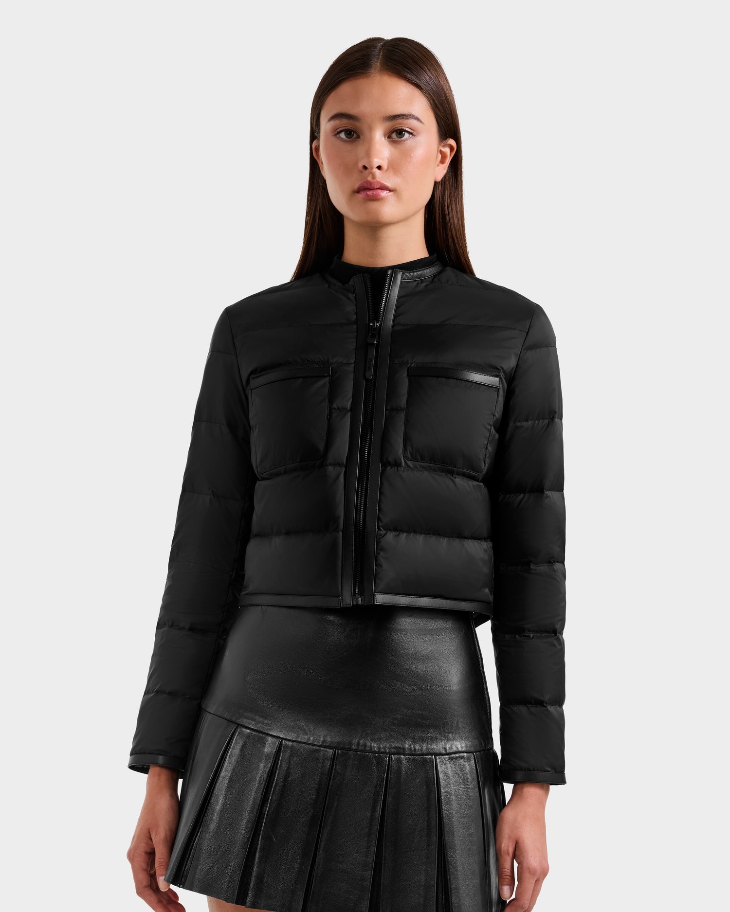 A woman wears the Rudsak MARKLE - 8125033 Black women’s cropped down jacket with a front zipper and chest pockets, styled with a black pleated leather mini skirt, standing against a plain light gray background.