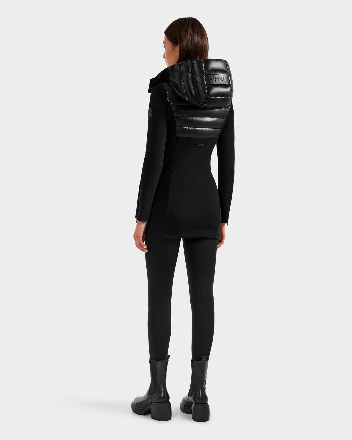 A woman faces away, wearing the Rudsak MARRY S - L125521 Black glossy shell jacket with a shiny padded hood, paired with black leggings and chunky weather-resistant boots against a plain light gray background.