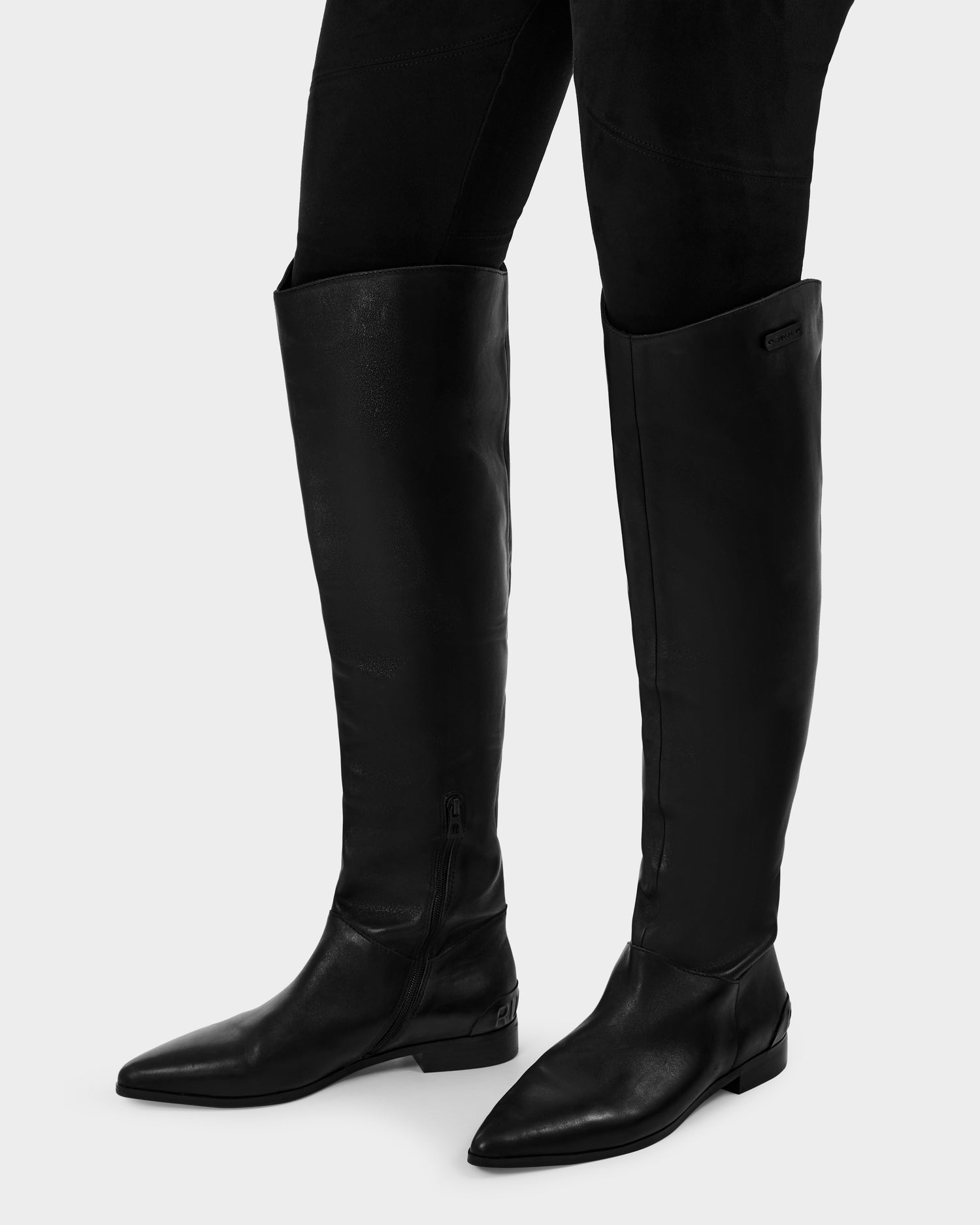 A person in black pants wears Rudsak's MARSALLA - 8225033 Black knee-high leather boots with pointed toes and low heels, standing against a light background. These sleek women’s boots complete the stylish look.