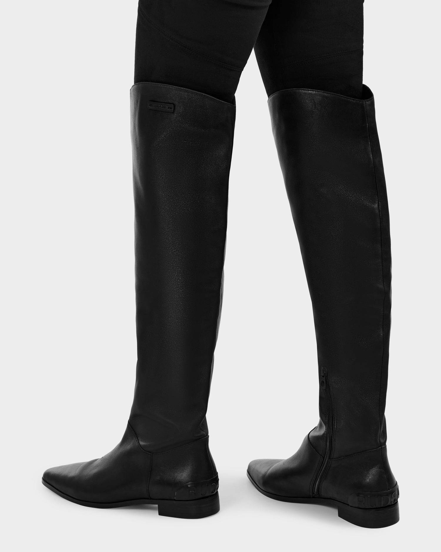A person wearing Rudsak MARSALLA - 8225033 Black smooth leather women’s high-shaft boots with a low heel and side zipper stands on a light gray background, showing only their legs and the boots.