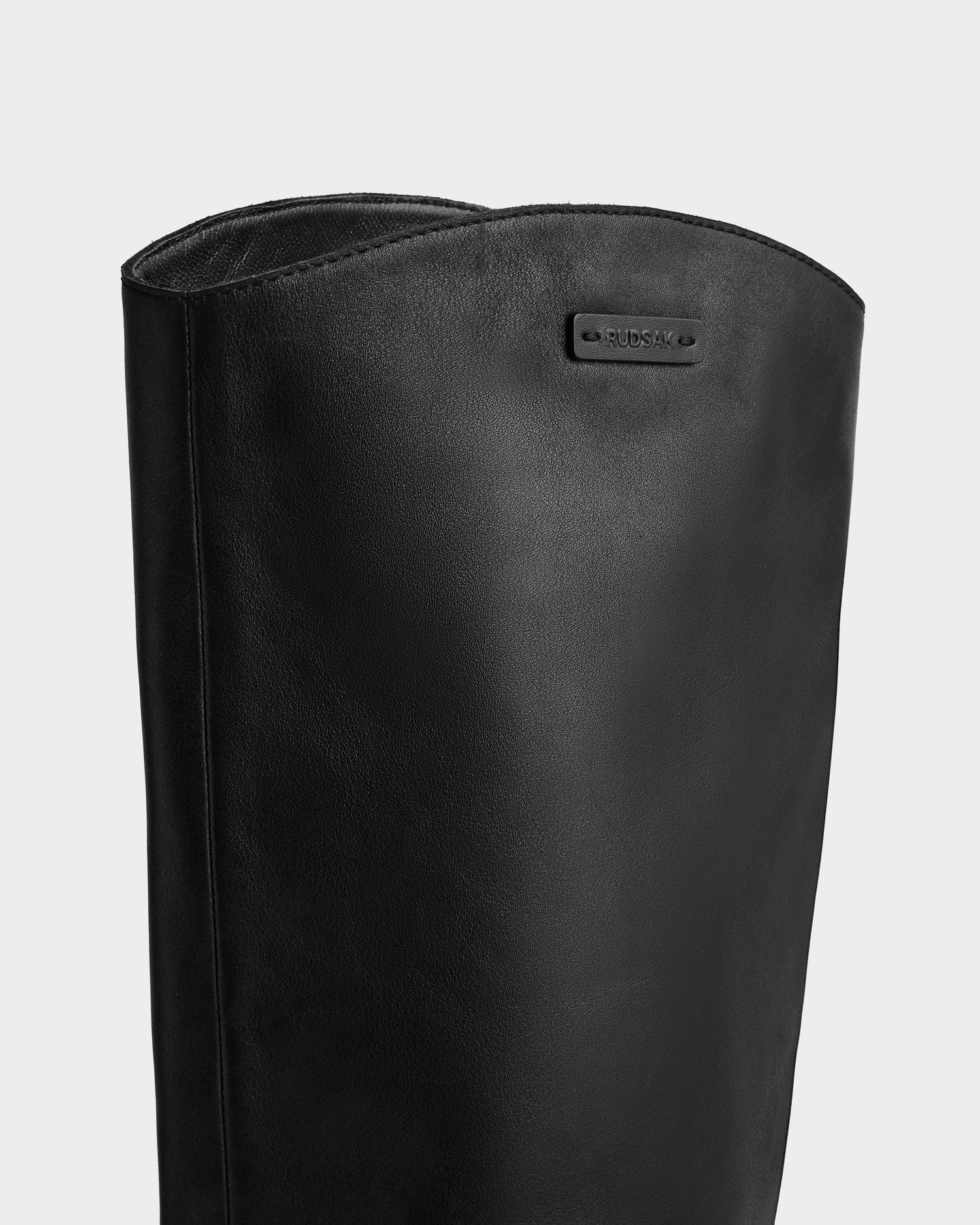 Close-up of the upper part of the MARSALLA - 8225033 Black women’s high-shaft leather boot by Rudsak, showcasing a small rectangular metal label with engraved text near the top edge against a plain light gray background.
