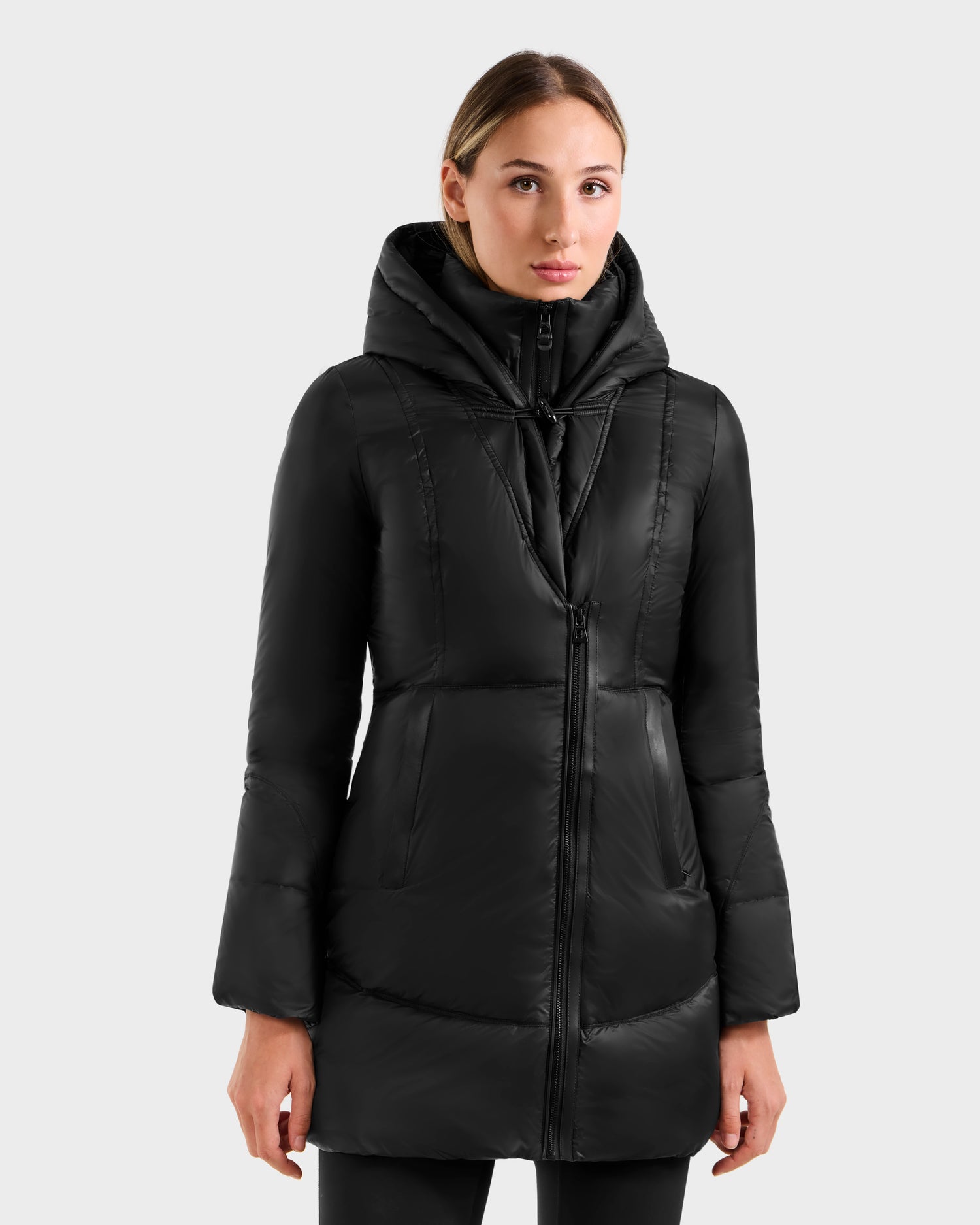 A woman wearing the Rudsak MARTINA - L125540 Black long quilted puffer coat with a removable fur-trimmed hood faces forward against a plain light grey background.