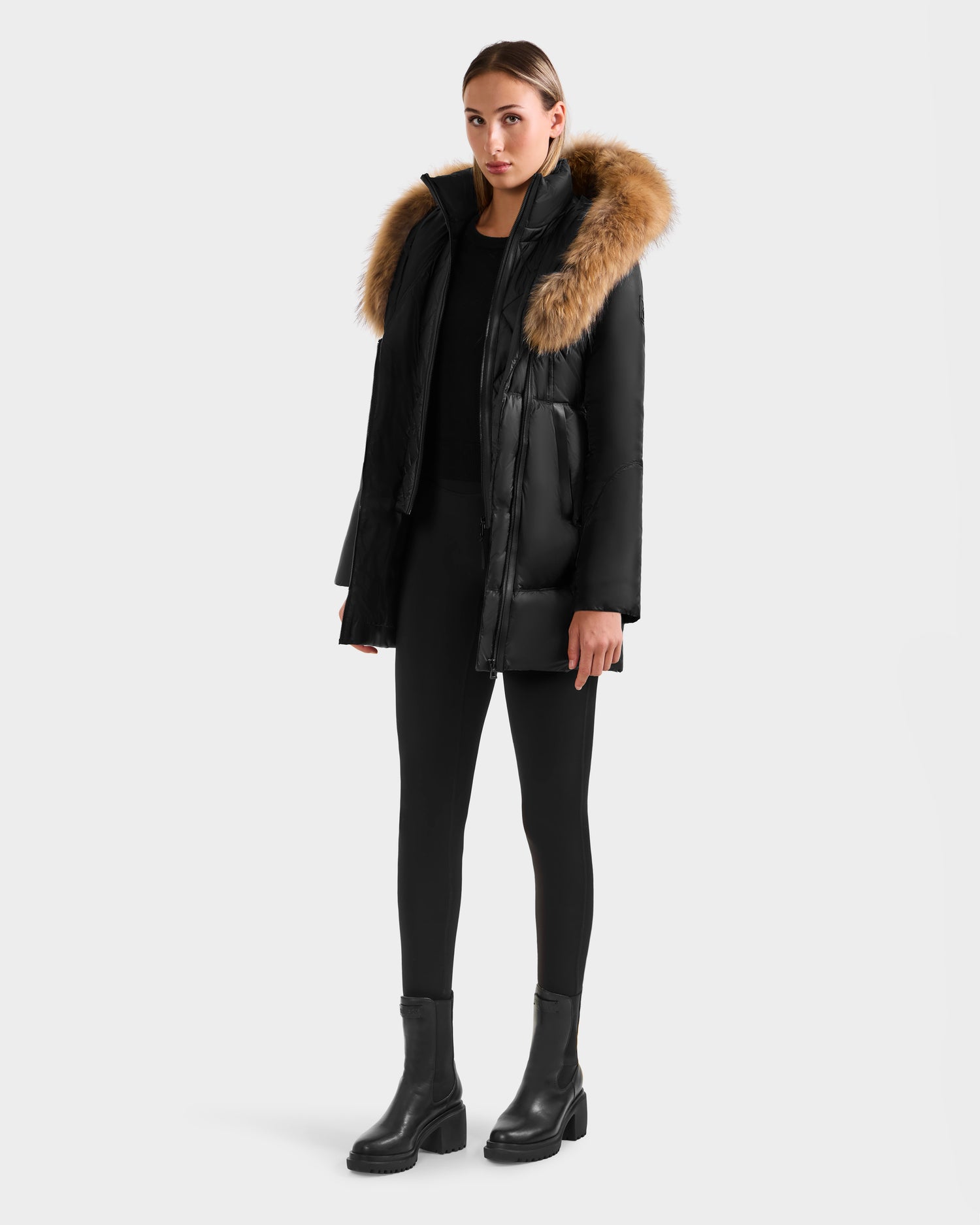 A person stands against a plain background wearing the Rudsak MARTINA - L125540 Black mid-length puffer jacket with removable fur-trimmed hood, paired with black leggings and black ankle boots.