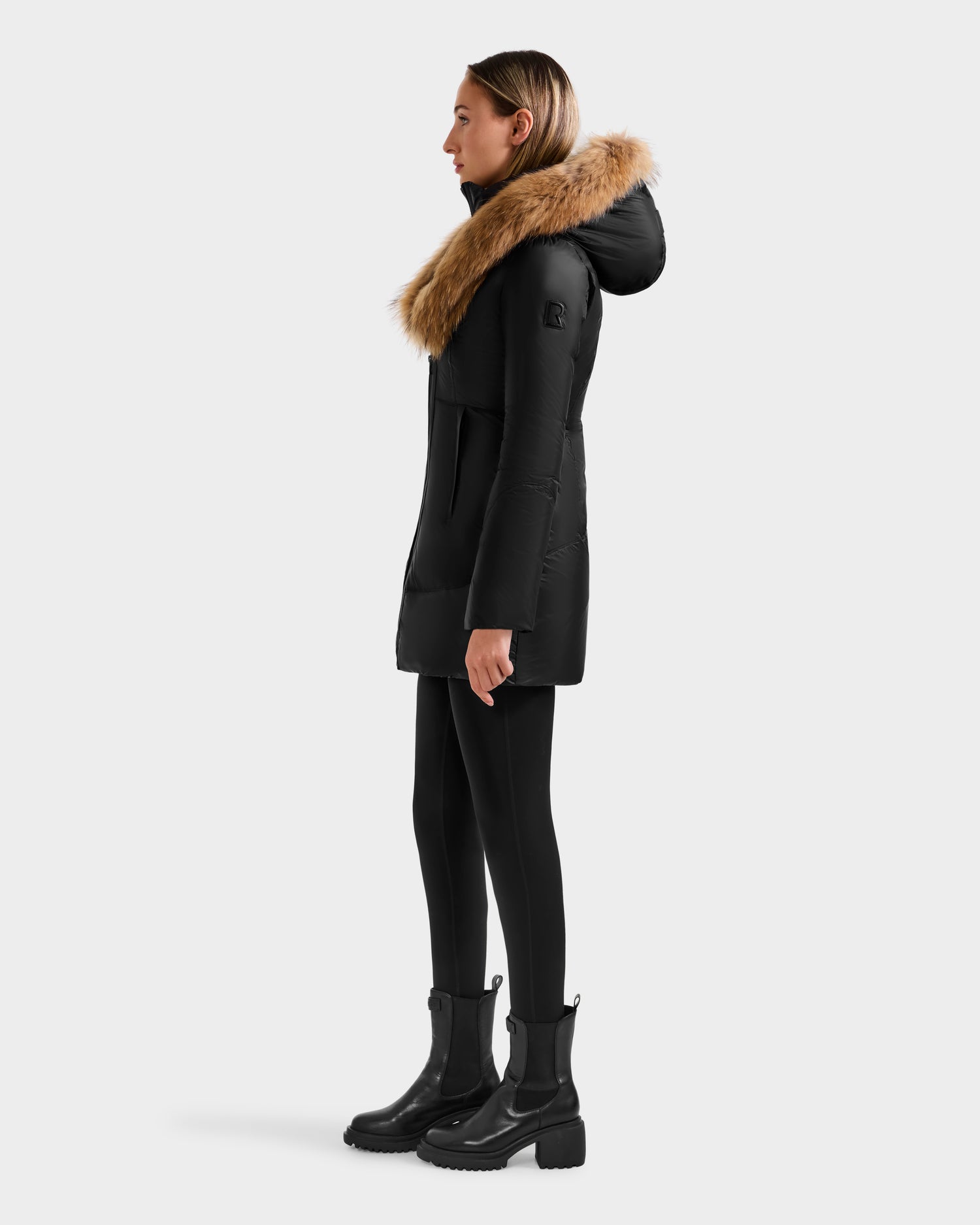 A woman stands in profile against a white background wearing the Rudsak MARTINA - L125540 Black mid-length puffer coat with a large removable fur-trimmed hood, paired with black leggings and black boots.