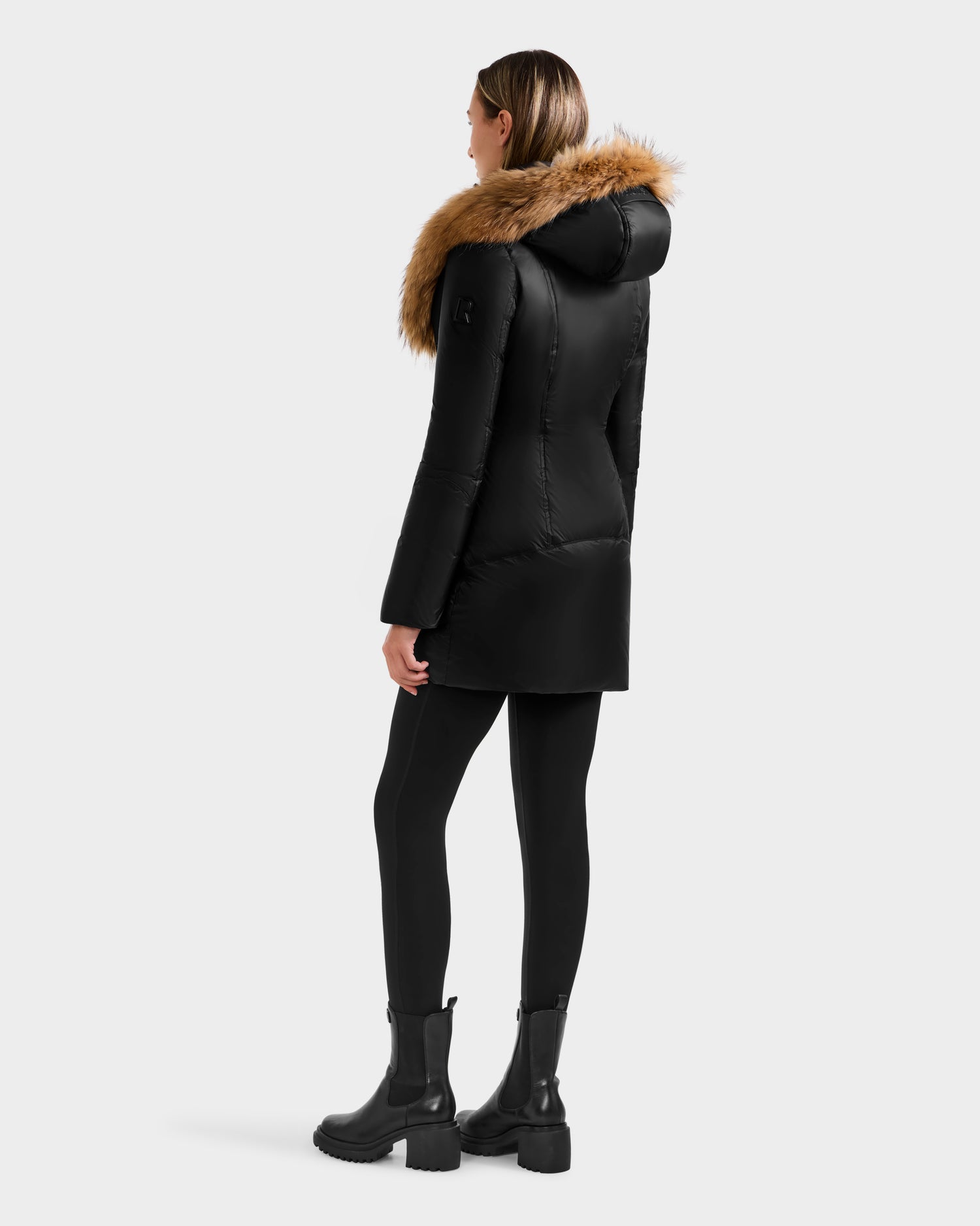 A person stands facing away in the Rudsak MARTINA - L125540 Black mid-length puffer coat with a large tan removable fur-trimmed hood, paired with black leggings and boots, set against a plain light background.