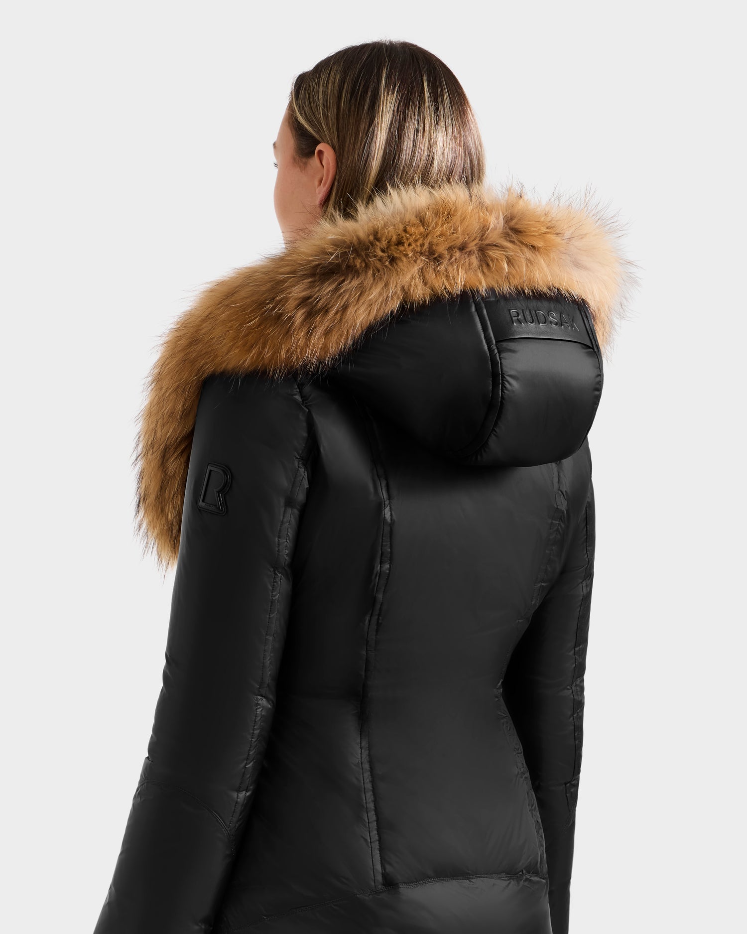 A woman with straight, light brown hair is seen from behind wearing the Rudsak MARTINA - L125540 Black mid-length puffer jacket with a large, fluffy removable fur hood border against a plain light gray background.