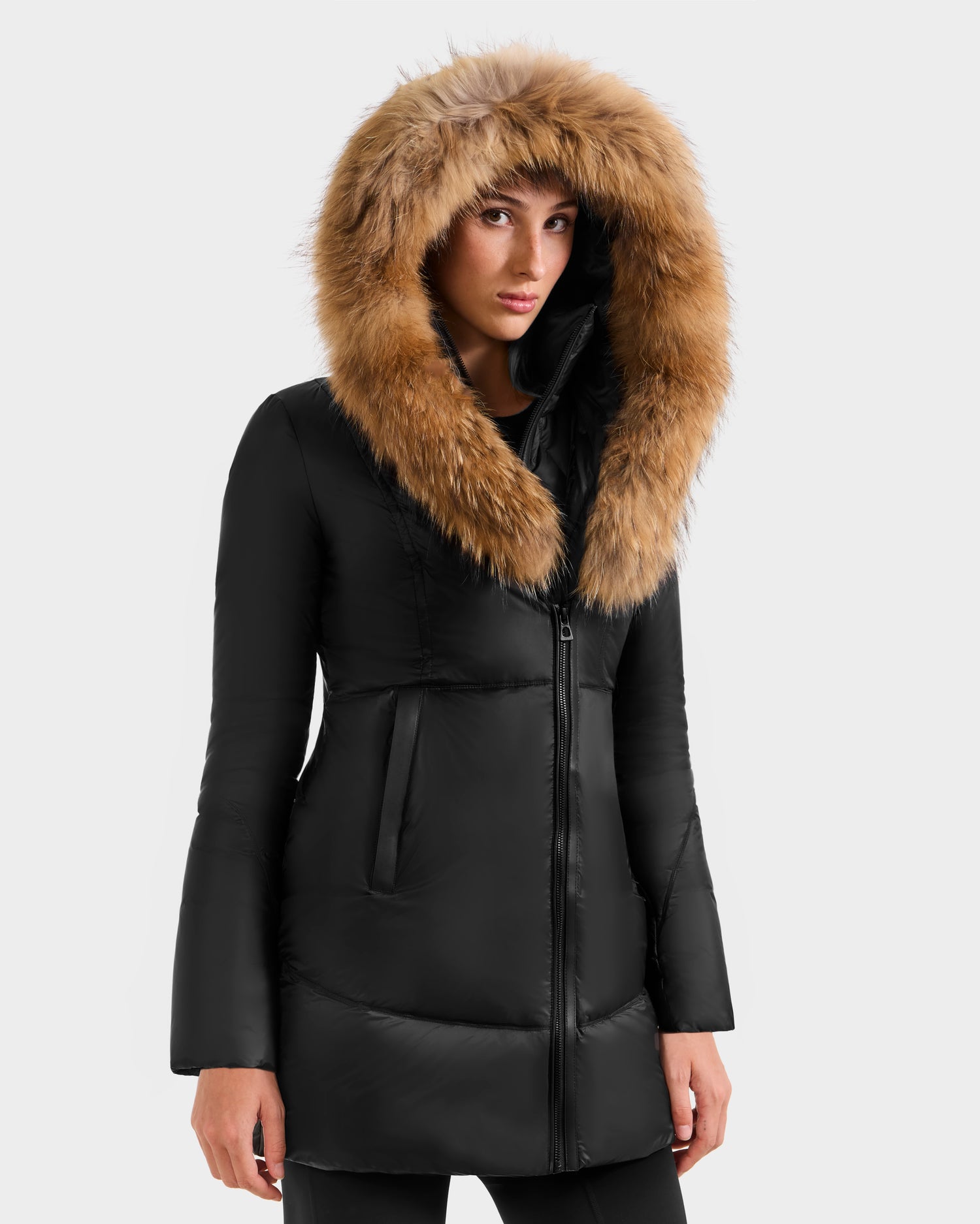 Someone stands facing forward against a plain light gray background, wearing the Rudsak MARTINA - L125540 Black mid-length puffer coat with a large, brown removable fur hood trim.
