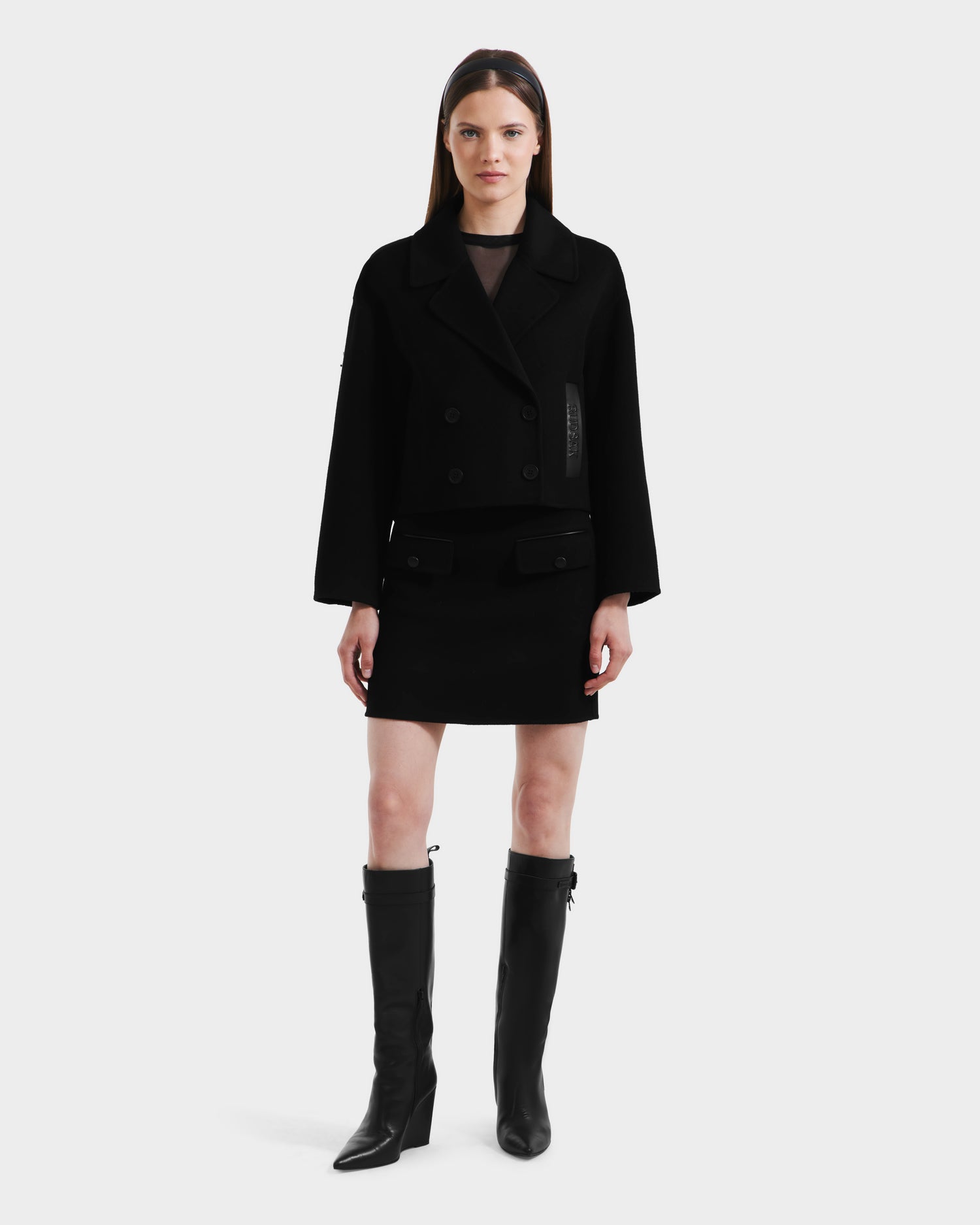 A woman stands facing forward, wearing the Rudsak MARTINEZ - 8125580 black vegan leather women’s short coat with a matching mini skirt and black knee-high boots against a plain white background.